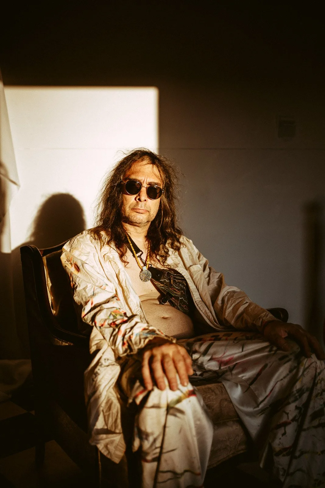 Ein Mann mit langem Haar, Sonnenbrille und legerer Kleidung sitzt auf einem Stuhl in einem Raum. Er trägt eine offene Jacke, eine Kette und seine Kleidung ist locker und auffällig.