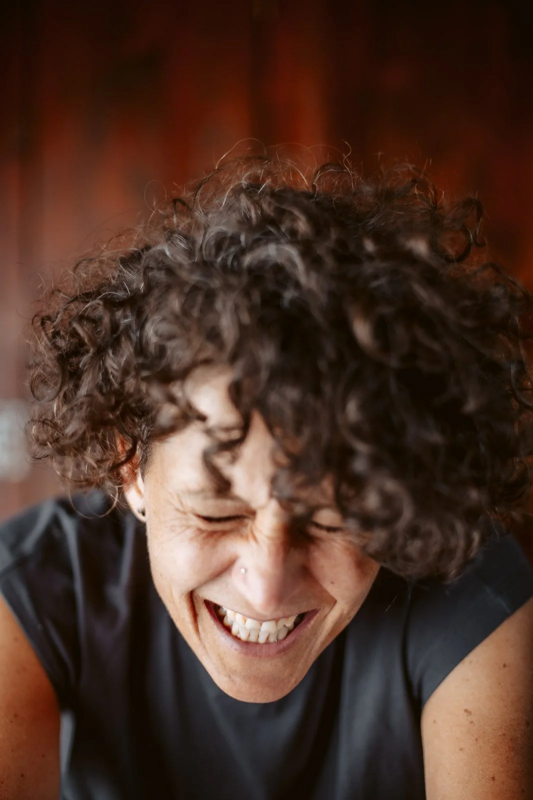 Eine lachende Frau mit lockigem Haar und Nasenpiercing, die sich offensichtlich amüsiert.