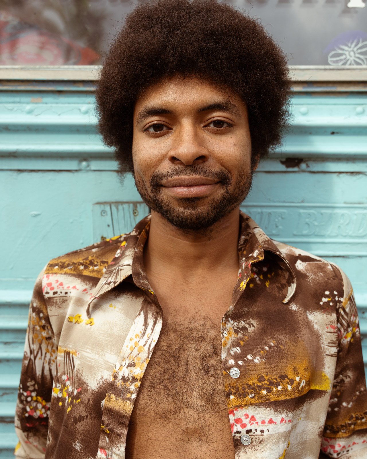 Ein junger Mann mit lockigem, braunem Haar und Bart trägt ein bunt gemustertes, ungeknöpftes Hemd. Er steht vor einer hellblauen Wand und lächelt leicht in die Kamera.