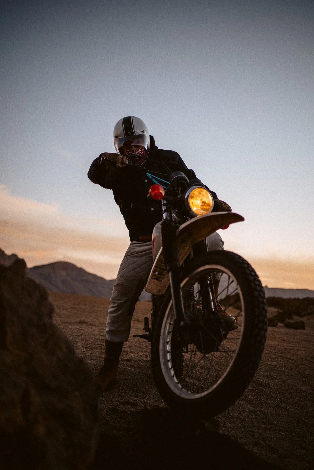 Ein Motorradfahrer trägt einen Helm und Motorradschutzkleidung, sitzt auf einem ausgestellten Offroad-Motorrad in einer Wüstenlandschaft bei Sonnenuntergang.