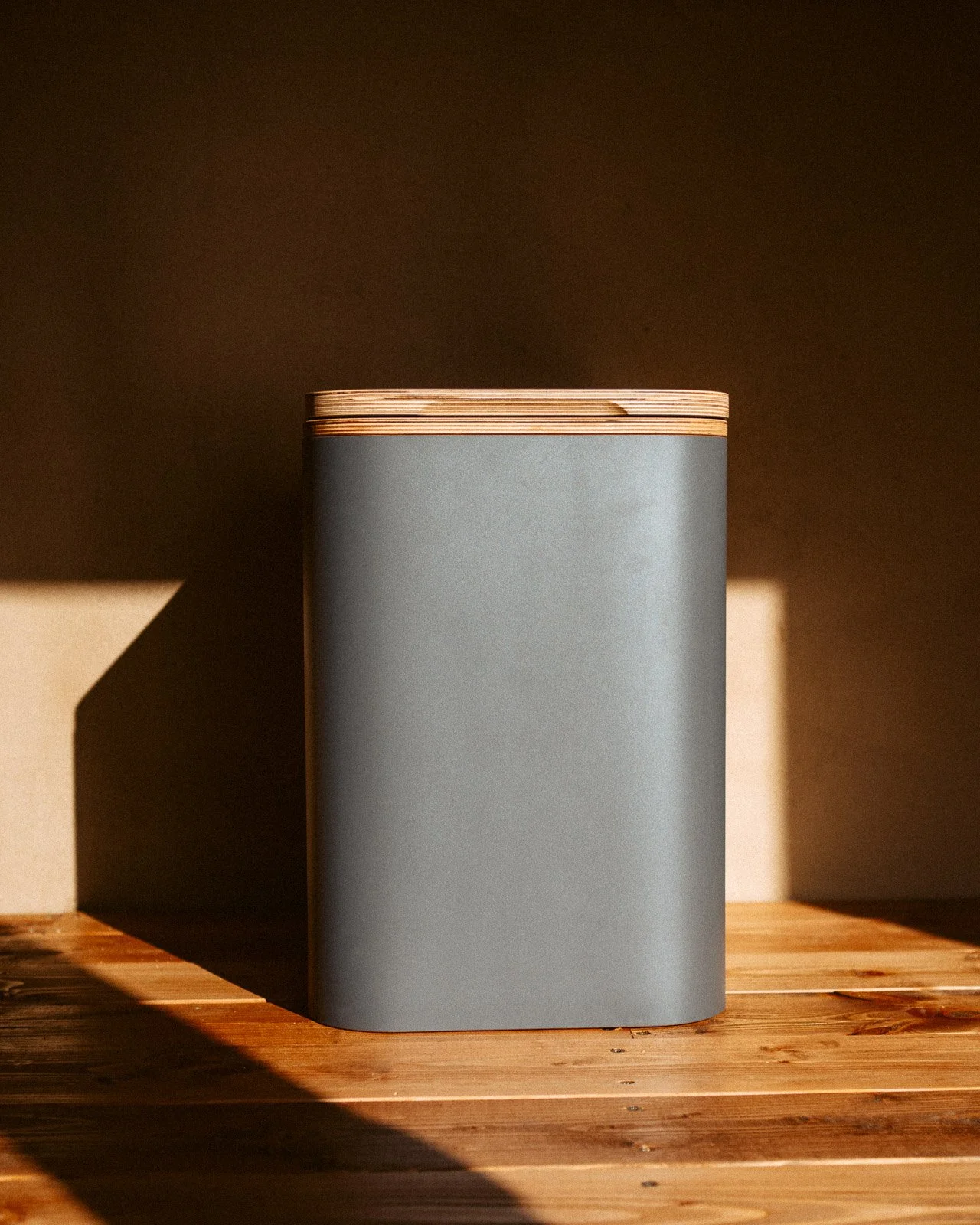 Ein grau-blauer rechteckiger Behälter mit holzartigem Deckel auf einem Holztablett, Licht- und Schatteneffekte.