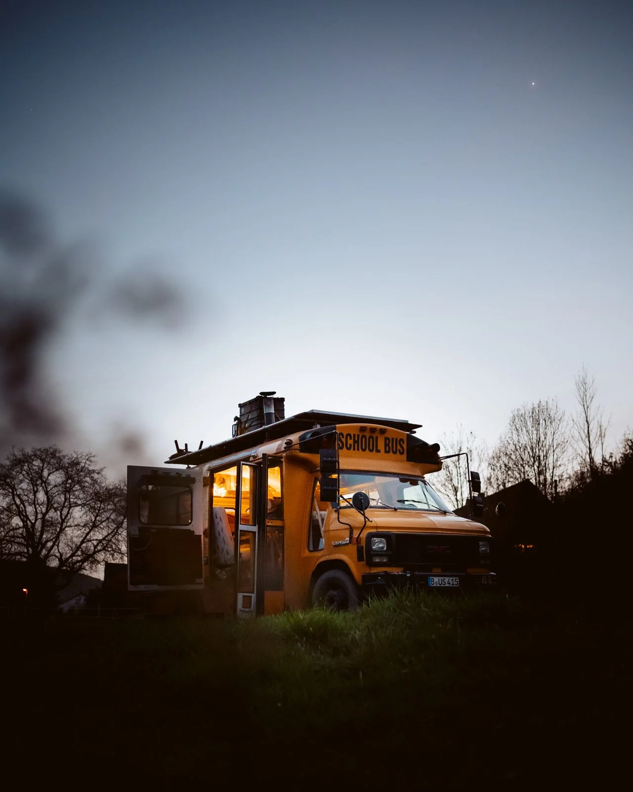Ein Schulbus steht nachts im Freien mit eingeschaltetem Licht, umgeben von dunklen Bäumen und einem dunklen Himmel.