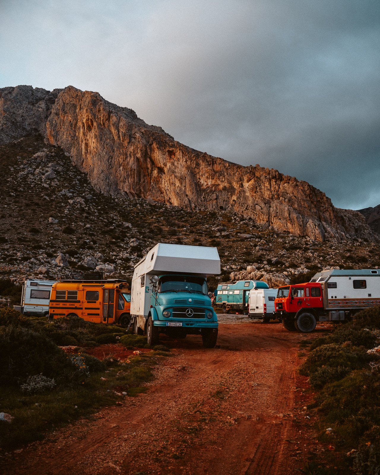 Campingfahrzeuge auf einem unbefestigten Weg vor einer Bergkulisse bei Sonnenuntergang.