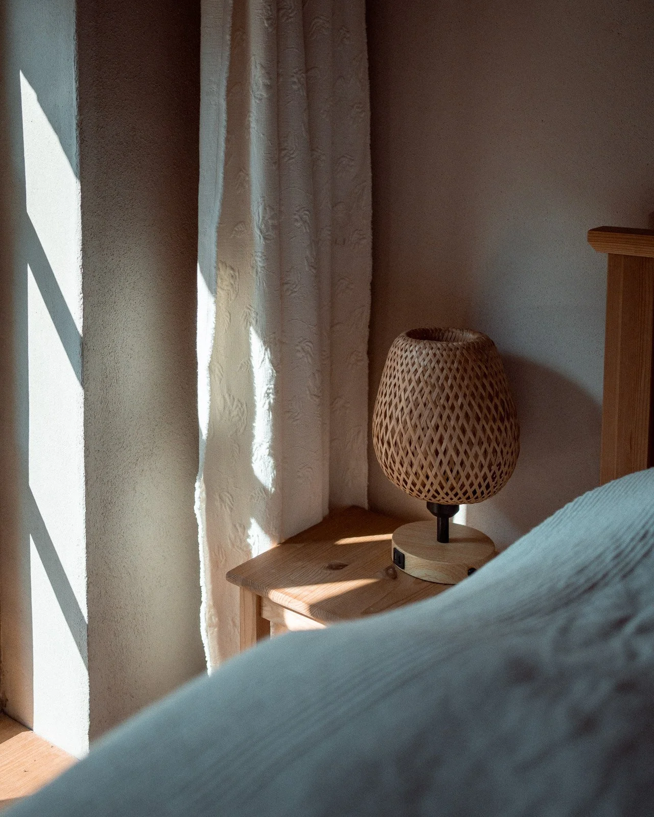 Ein Nachttisch mit einem Lampenschirm aus geflochtenem Material, neben einem Bett mit weißer Bettwäsche, vor einem Fenster mit weißen Vorhängen, durch das Sonnenlicht fällt.