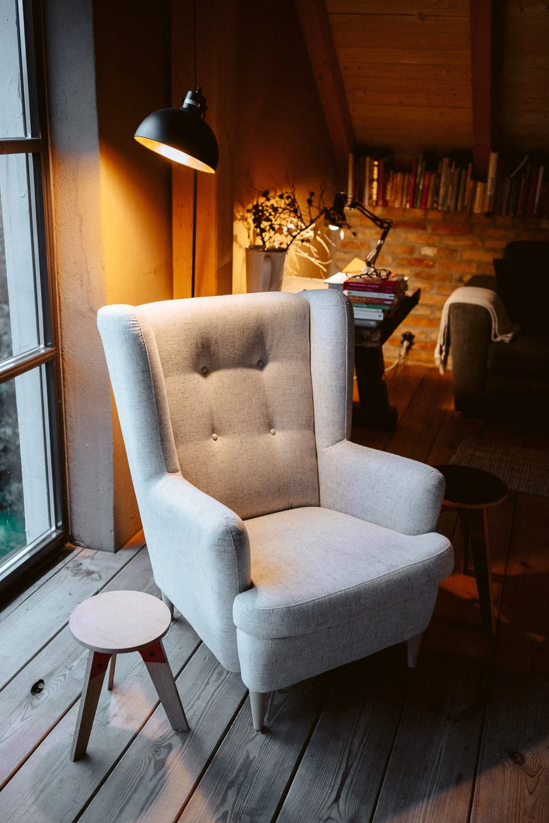Gemütliches Wohnzimmer mit einem hohen, gepolsterten Sessel in Hellgrau neben einem kleinen Holztisch. Im Hintergrund stehen Bücherregale, ein Tisch mit Büchern und eine Stehlampe, beleuchtet mit warmem Licht, in einem Raum mit Holzdecke und Holzfußb