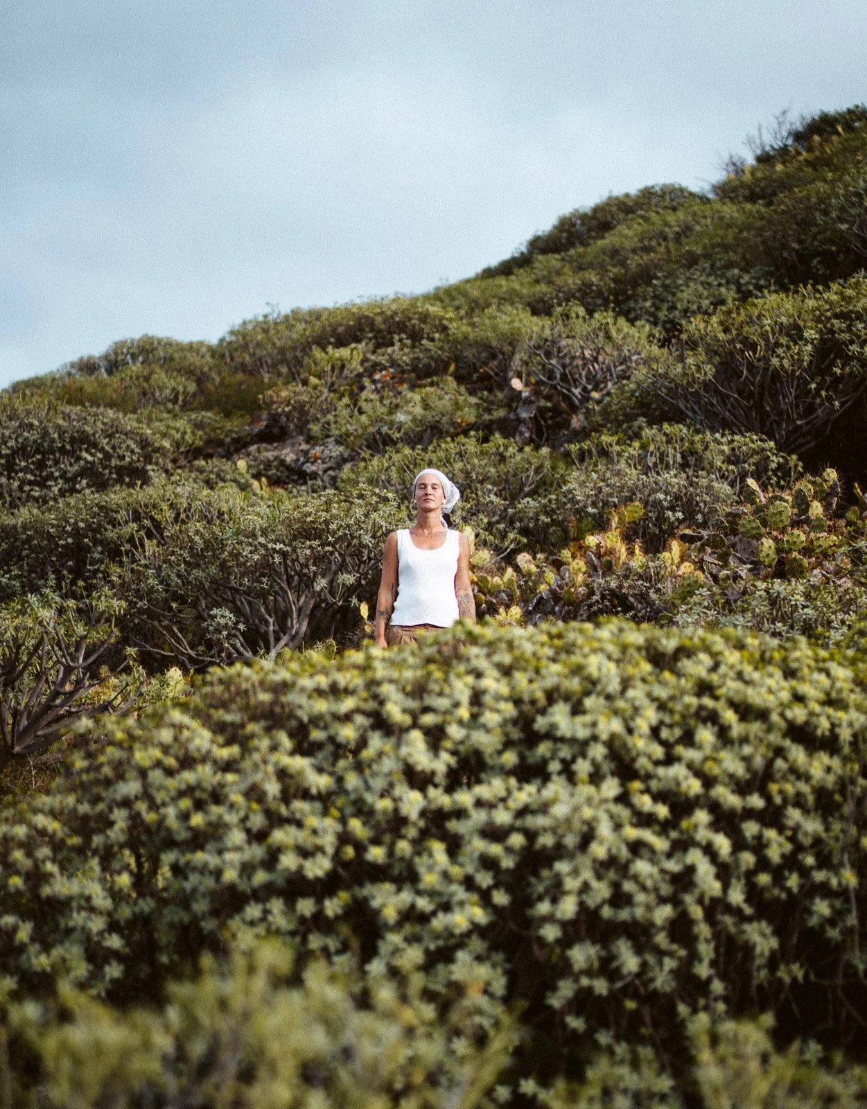 Frau in weißer ärmelloser Bluse steht inmitten von Sträuchern und Vegetation, im Hintergrund Himmel.