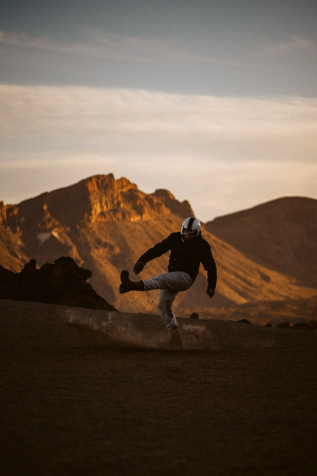 Ein Person beim Kickflip auf einem Motorrad in einer Wüste bei Sonnenuntergang, im Hintergrund Berge.