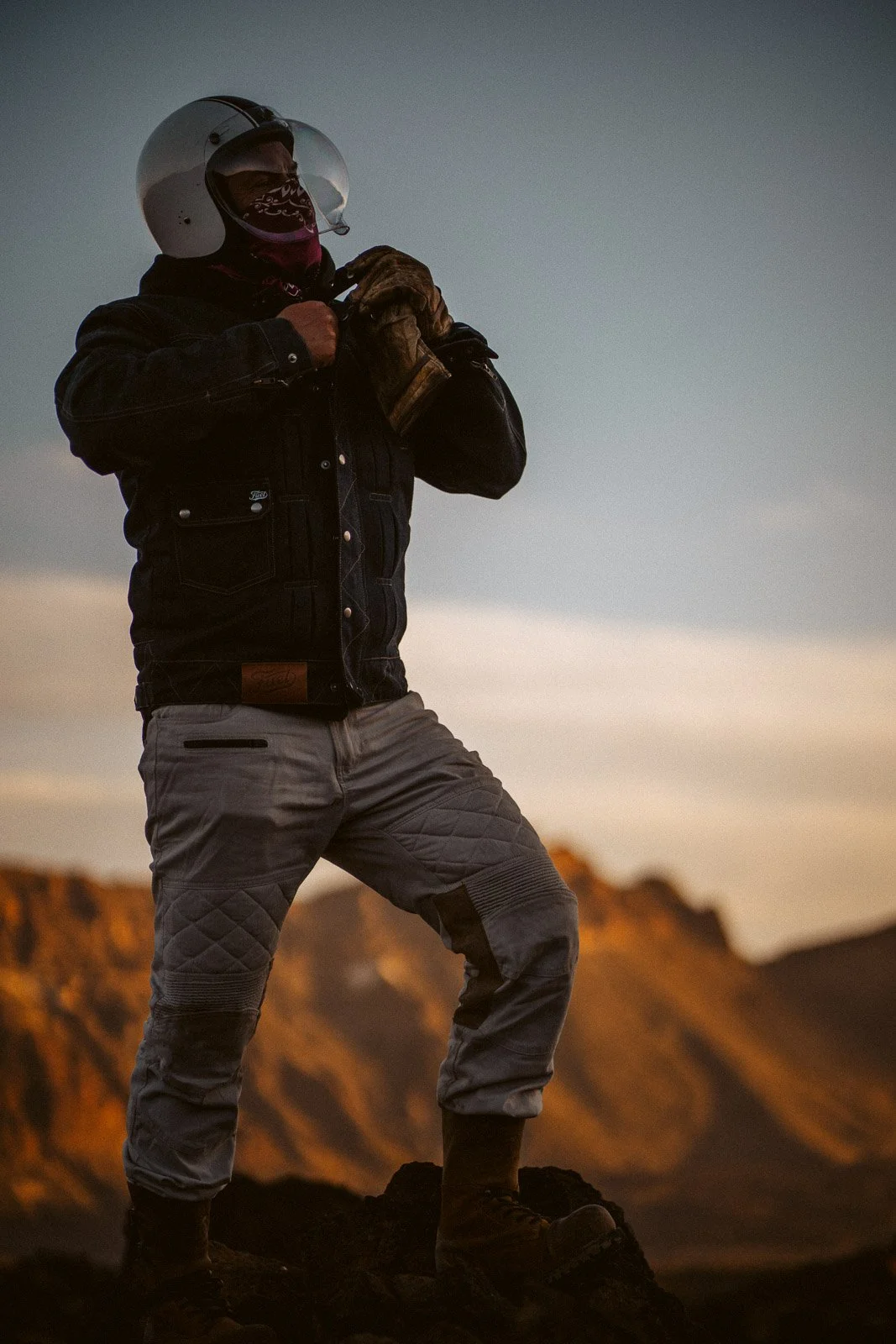 Ein Mann steht in einer Berglandschaft bei Sonnenuntergang, trägt eine Motorradjacke, eine Motorradhelm und Kletterstiefel, hält einen Handschuh in der Hand.