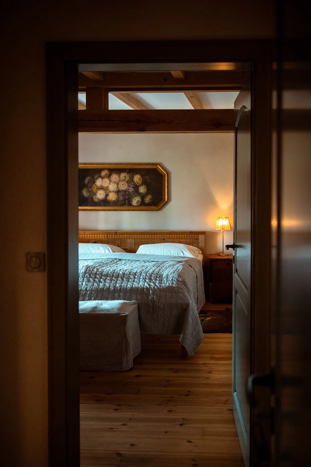 Ein gemütliches Schlafzimmer mit einem Bett, zwei weißen Kissen und einer Quiltvorderseite. Ein Nachttisch mit einer Lampe befindet sich rechts neben dem Bett. An der Wand hängt ein Blumenbild in einem goldenen Rahmen.