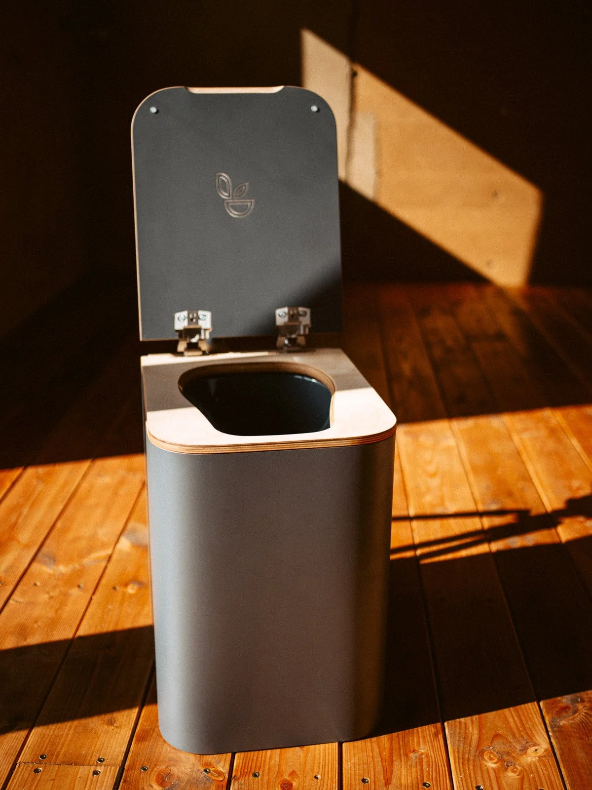 Ein moderner Mülleimer aus Metall mit einem offenen Deckel, auf einem Holztisch, im Hintergrund eine Wand und ein Schatten.