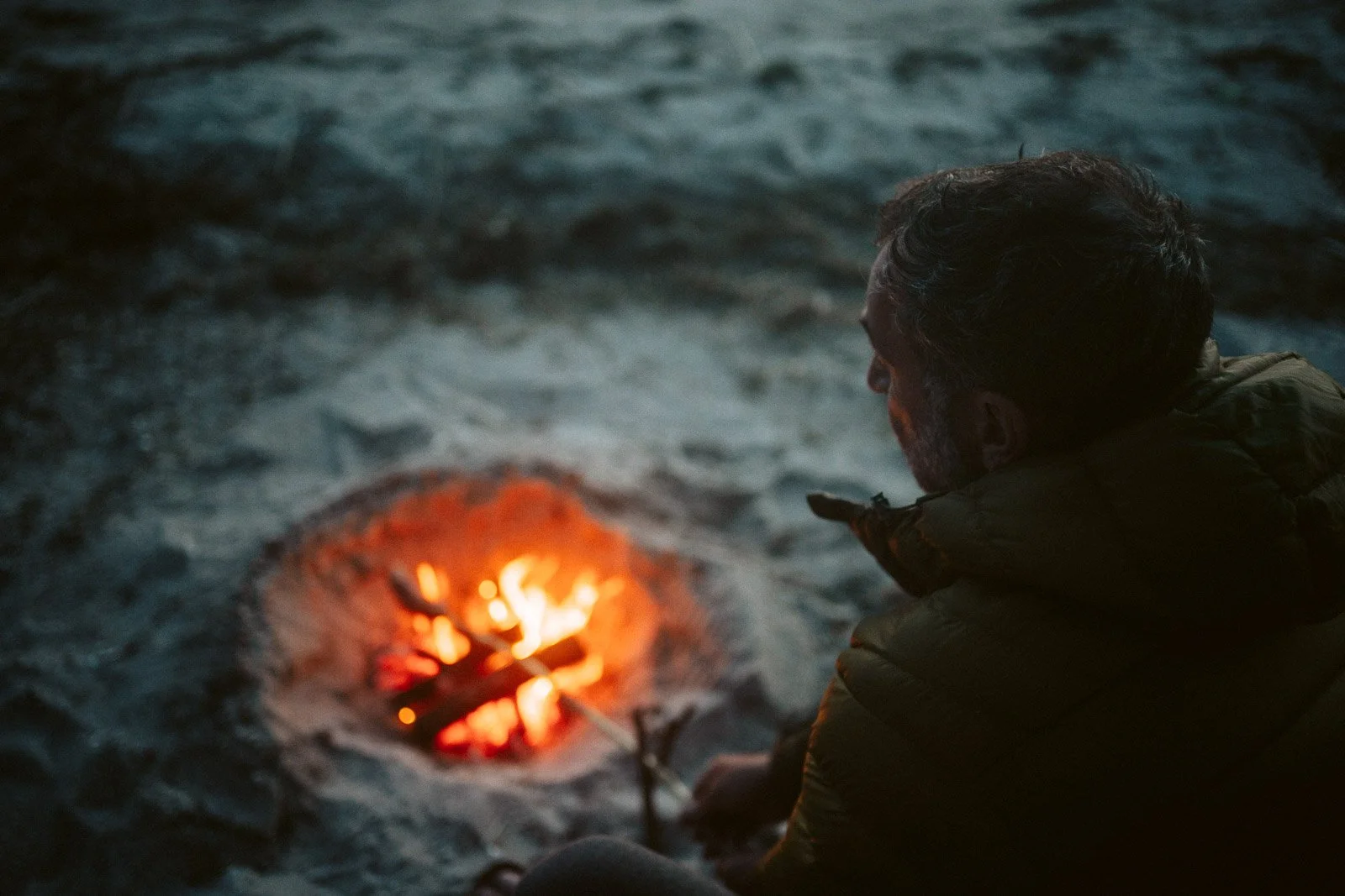 Ein Mann sitzt am Strand in der Dunkelheit und blickt auf ein kleines Feuer, das im Sand brennt.