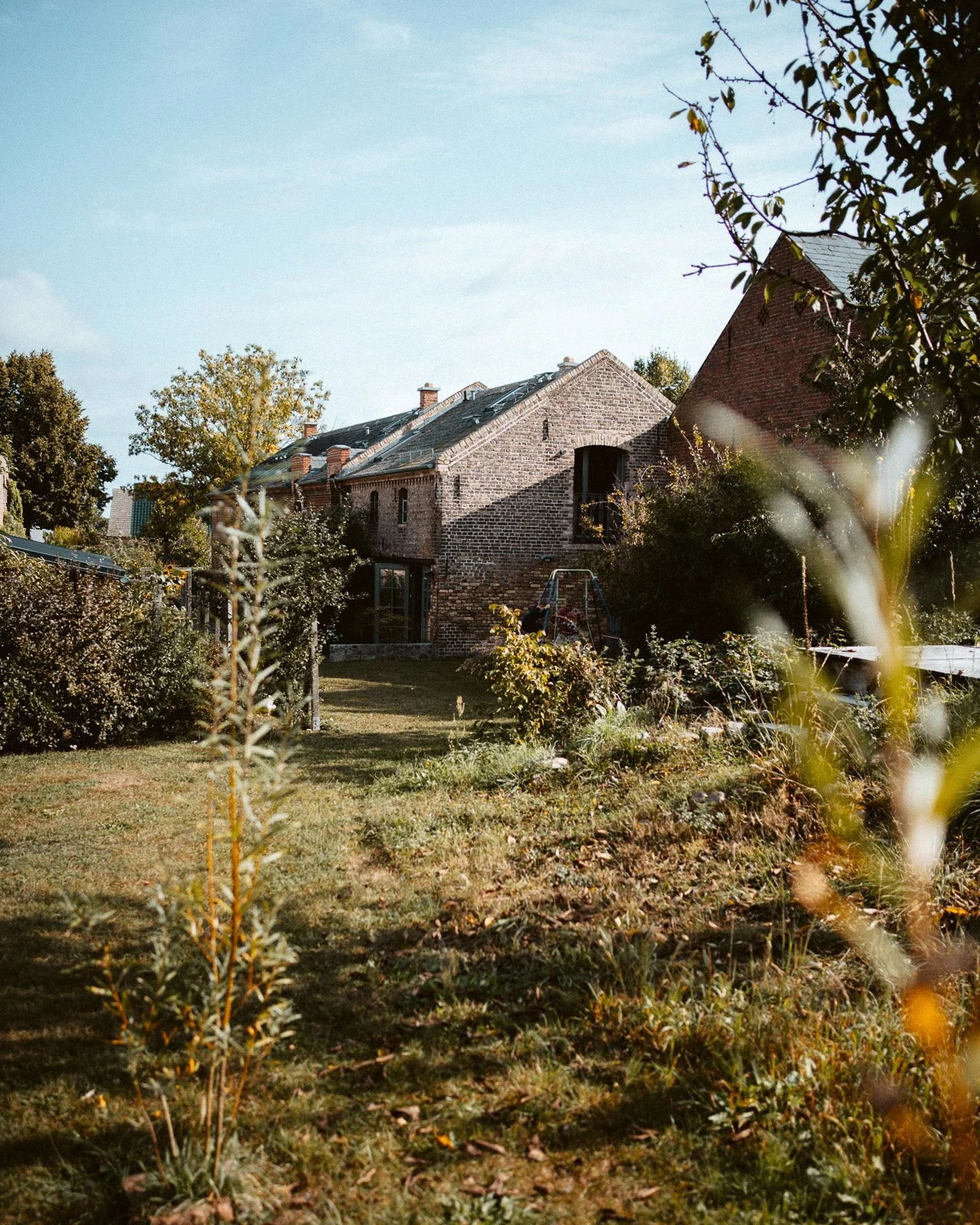 Ein Garten mit einem gepflegten Rasen, Büschen und Bäumen. Im Hintergrund sieht man ein Gebäude aus roten Ziegelsteinen mit mehreren Fenstern und eine Schaukel im Garten.