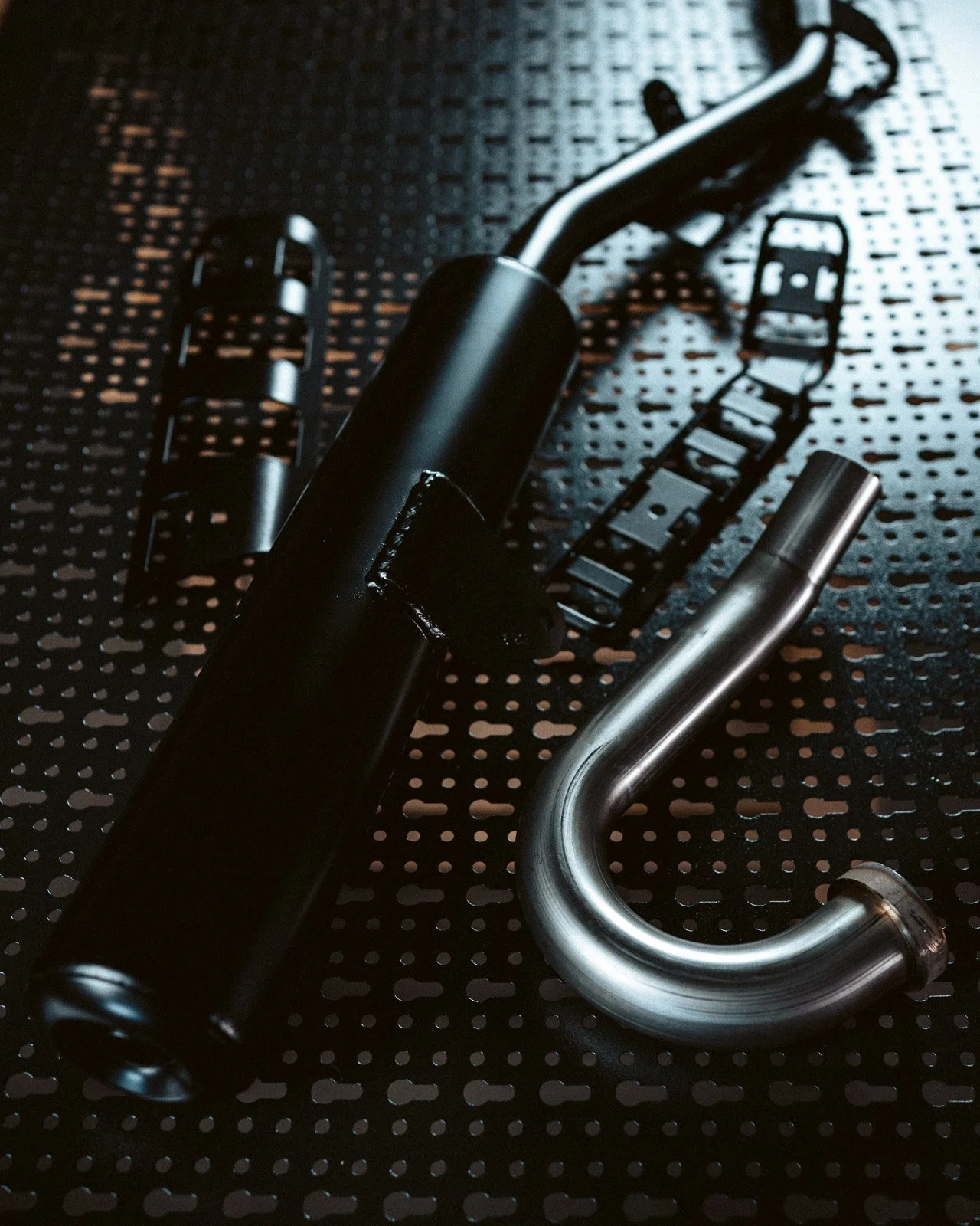 Drei Motorradteile, inklusive Stoßdämpfer, Auspuffrohr und Kettenschutz, liegen auf einer perforierten schwarzen Oberfläche.