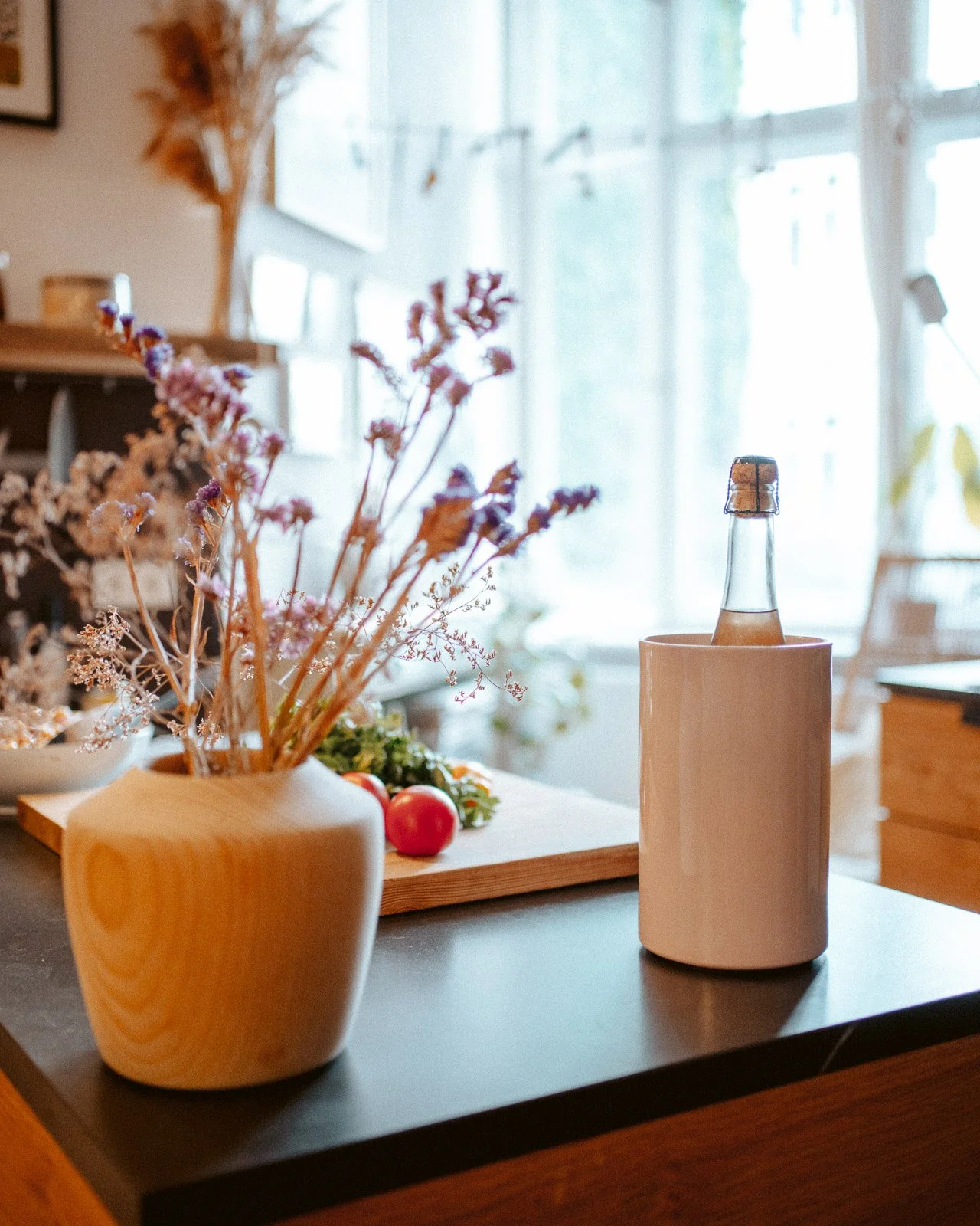 Bunte Trockenblumen in einer Holze Vase, frische Tomaten auf einem Schneidebrett, eine Flasche mit Getränk in einem weißen Keramikhalter, in einem hellen, gemütlichen Raum mit großen Fenstern.