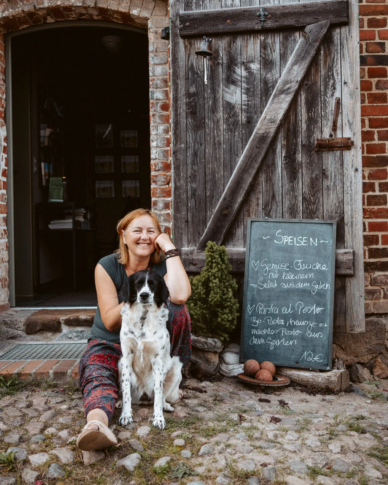 Frau sitzt draußen mit Hund vor einem alten Holzverschlag, neben einem P-Schild mit Speisen und Preisen, bei einem kleinen Baum und einem Steinplattenweg.