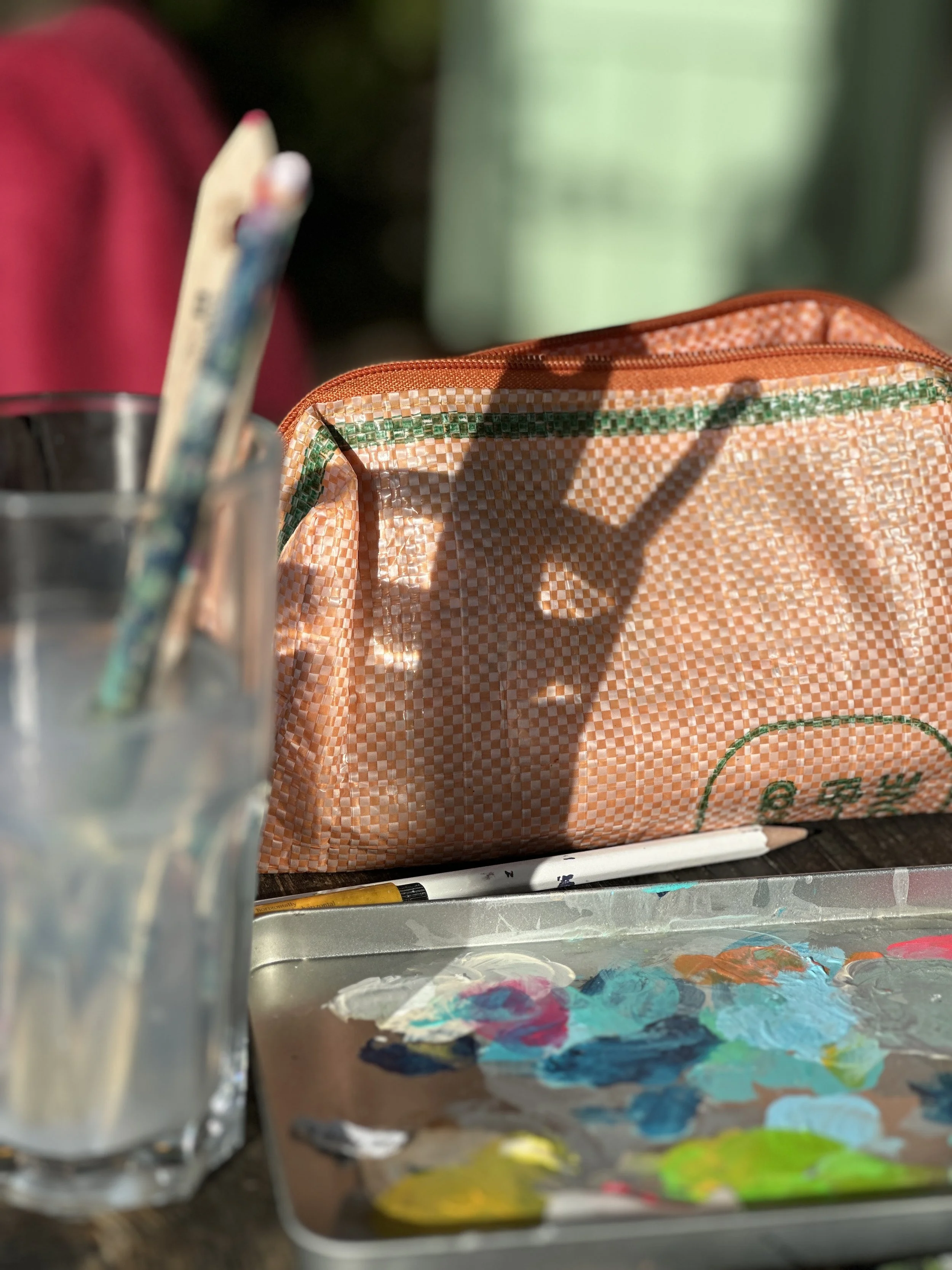 Gelbe und grüne Acrylfarben auf einer Farbpalette, eine zerknautschte orangefarbene Tasche mit grünen Streifen, Pinsel in einem Glas.