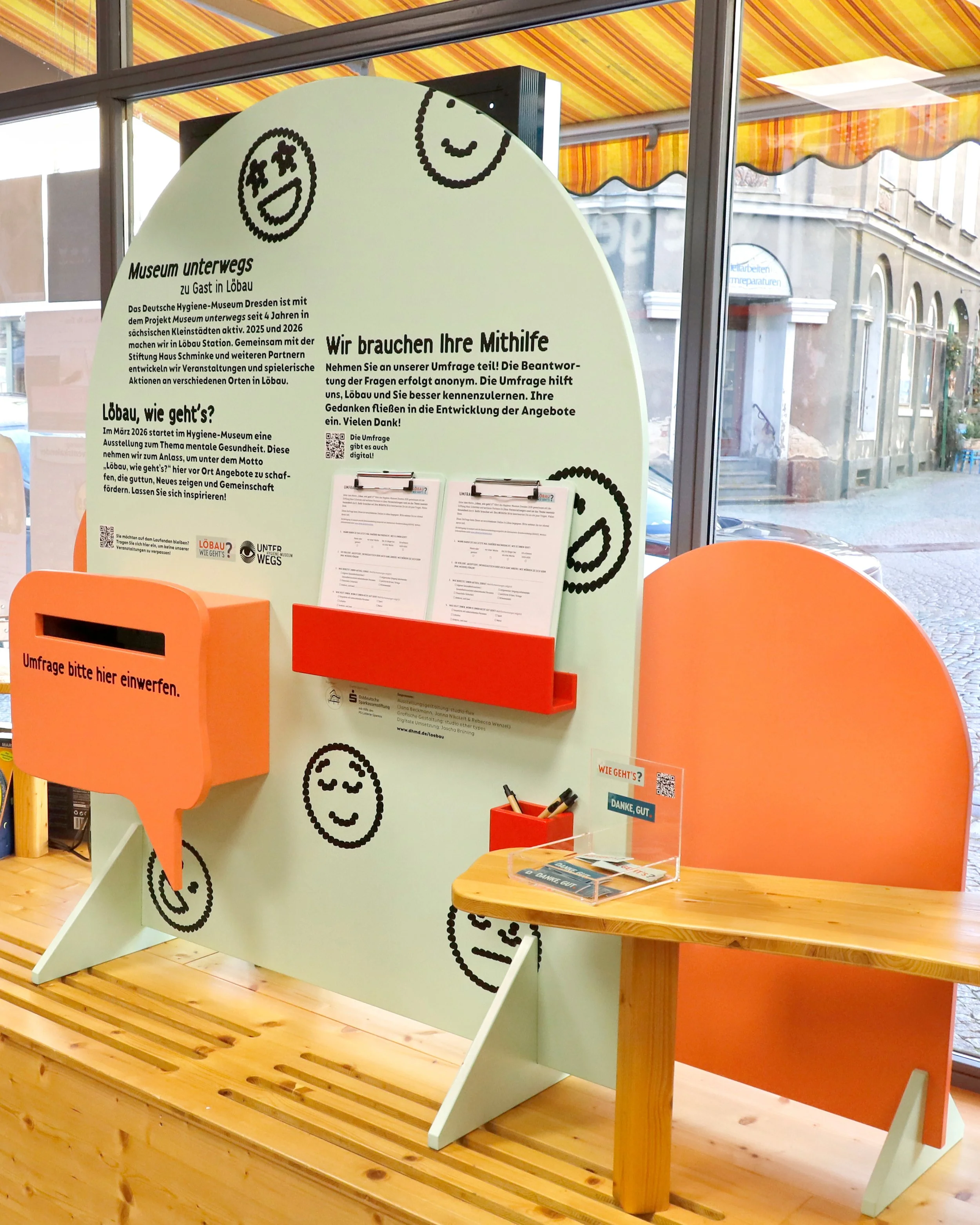 Befragungsstation mit orangefarbenem Einwurfkasten, Umfragebögen auf Klemmbrettern und Holztisch vor Fenster mit gelb-orange gestreiftem Markisendach