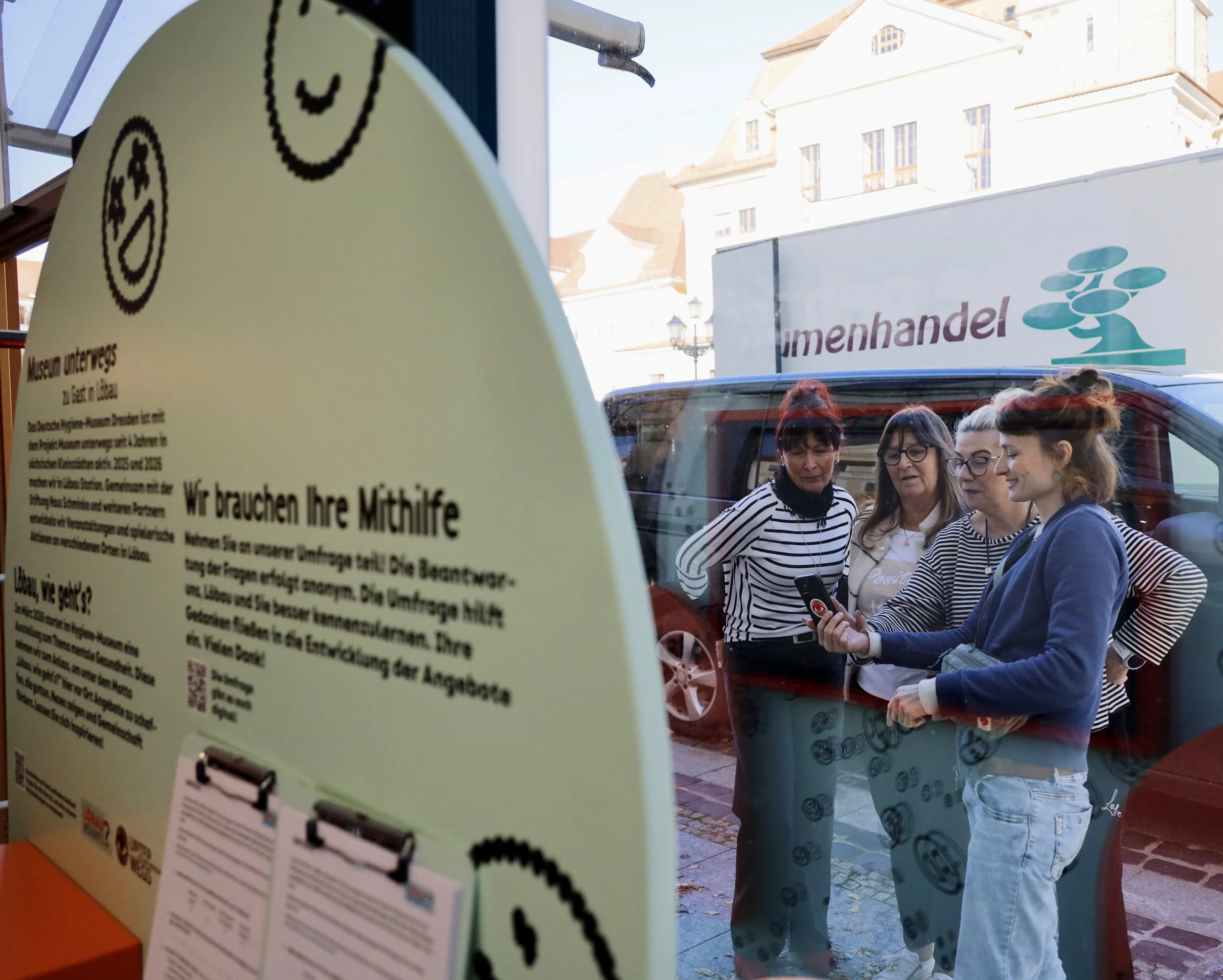 Vier Frauen stehen vor einer Glaswand mit Text und halten ein Smartphone in der Hand