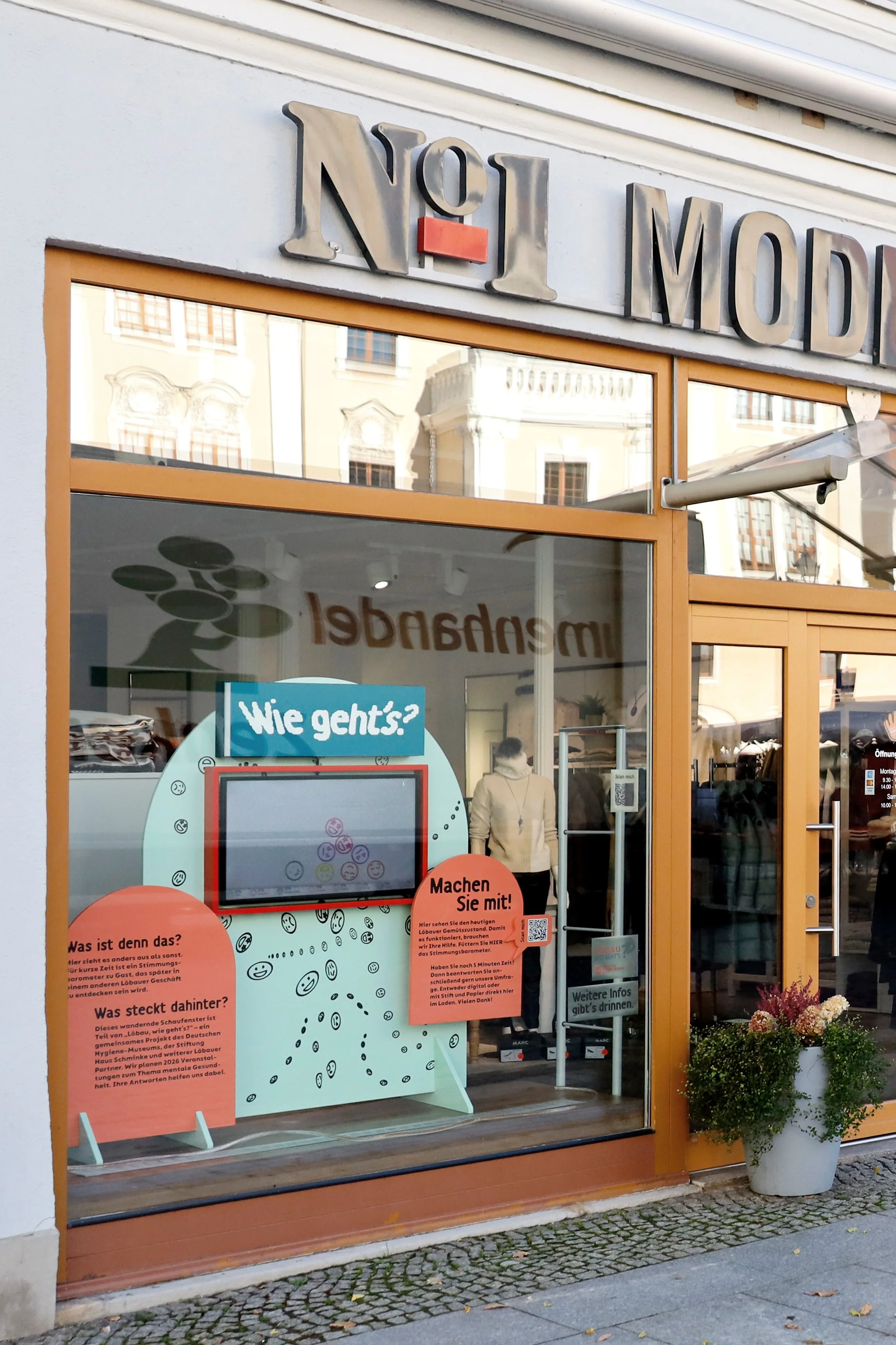 Schaufenster mit interaktivem Stimmungsbarometer und zwei orangefarbenen Informationstafeln mit Texten zu einem Projekt namens »Löbau, wie geht's?«