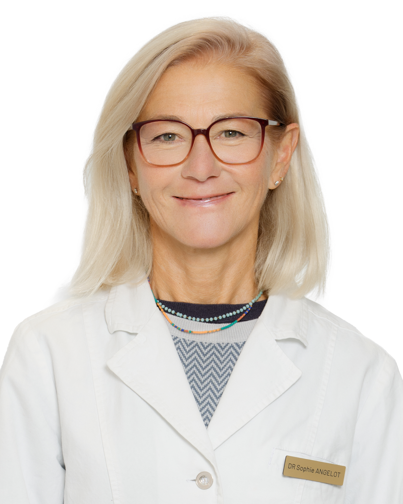 Une femme blonde portant des lunettes, un uniforme blanc, une veste avec un badge, souriante, dans un cadre professionnel.
