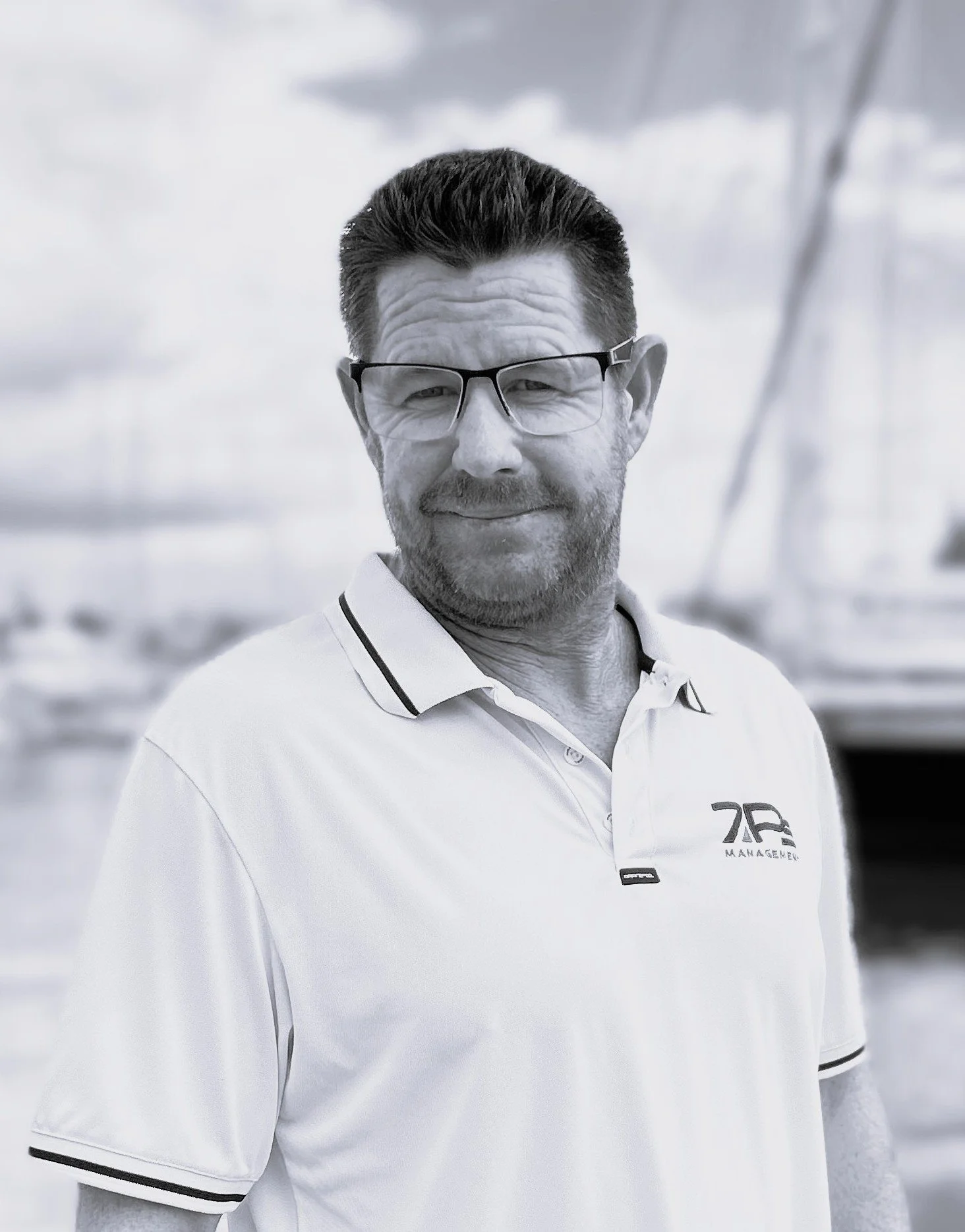 A man with dark hair, glasses, and a beard, wearing a white polo shirt with black trim, standing outdoors with boats in the background, black and white photograph.