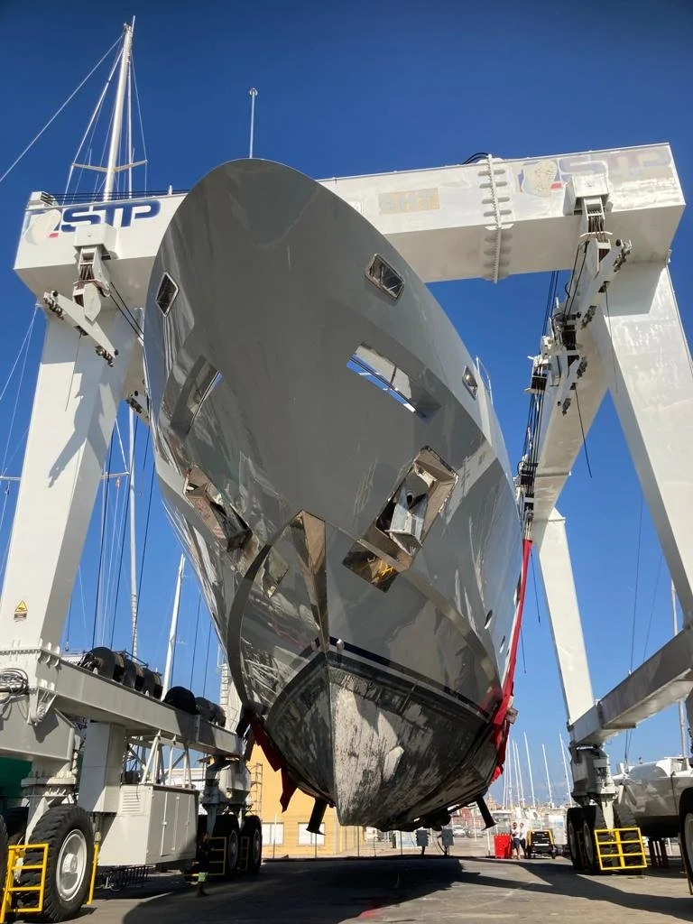 A large yacht is suspended in the air by a boat lift at a marina, with a clear blue sky in the background.