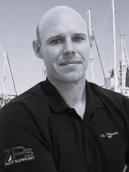 A man with a shaved head wearing a black polo shirt with yacht support logos, standing outdoors with boats and masts visible in the background.