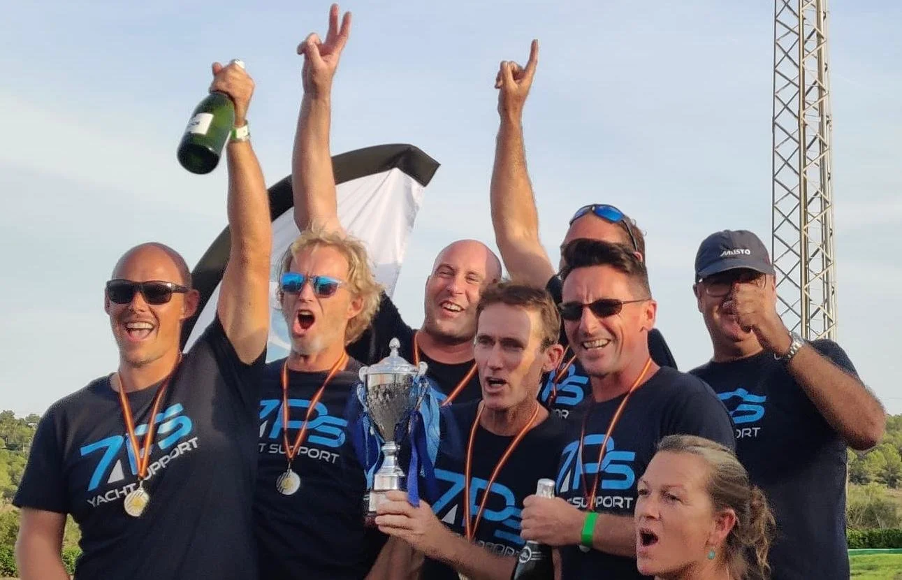 Group of six men and one woman celebrating on a sports field, holding a trophy, medals, and champagne, wearing matching black shirts with blue lettering, some with sunglasses, with a clear sky and a metal tower in the background.