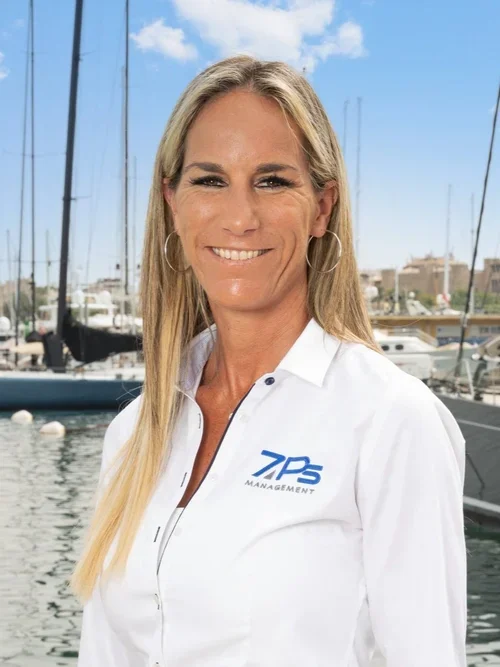 A woman with long blonde hair wearing a white collared shirt with the 7 Peaks Management logo, standing in front of a marina with boats and a partly cloudy sky.