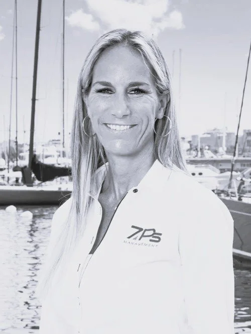 A woman with long hair wearing a white jacket with a logo, standing in front of a marina with sailboats, smiling at the camera.