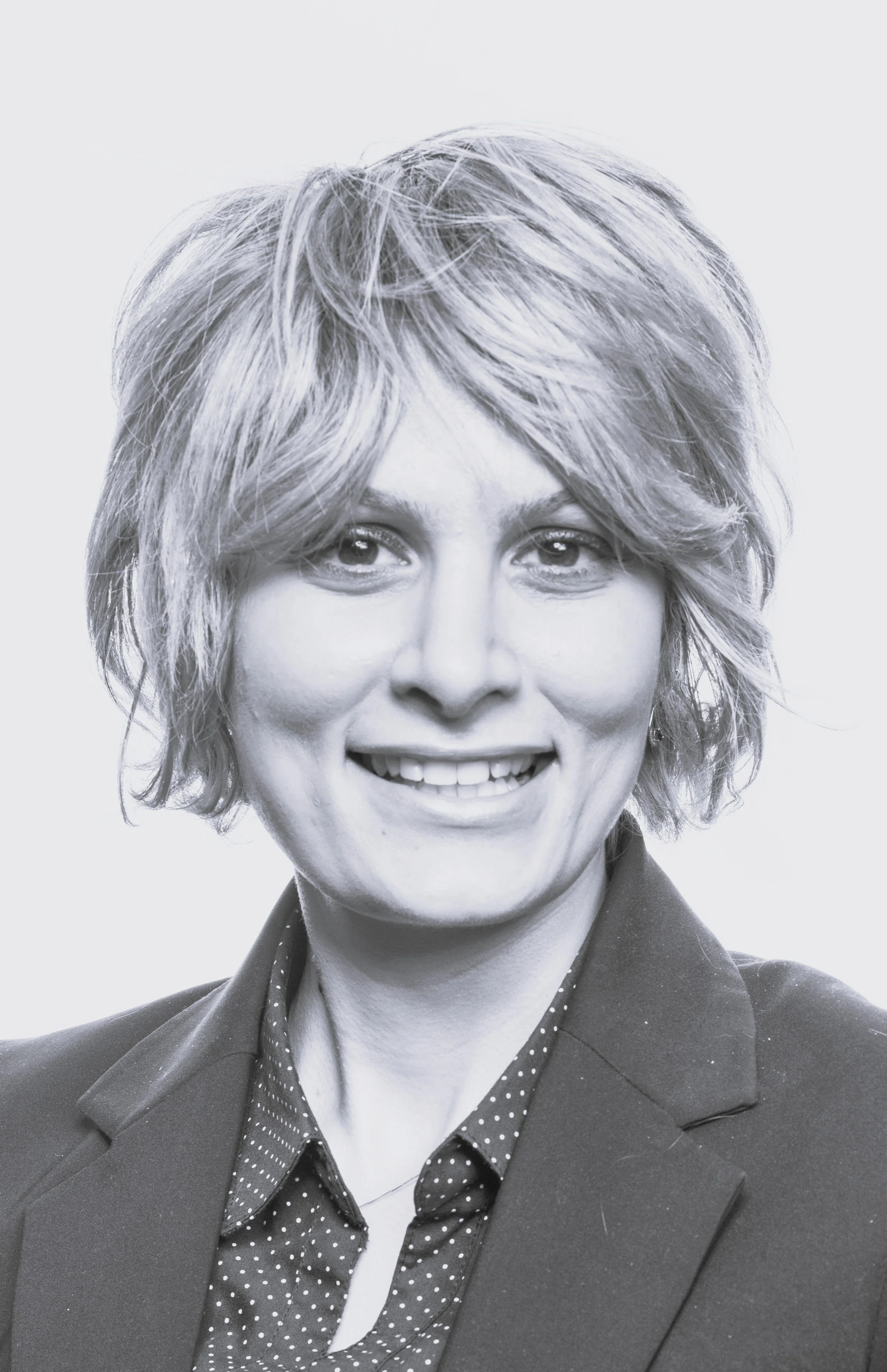 Black and white portrait of a woman with short, wavy hair, smiling, wearing a blazer over a polka-dotted shirt.