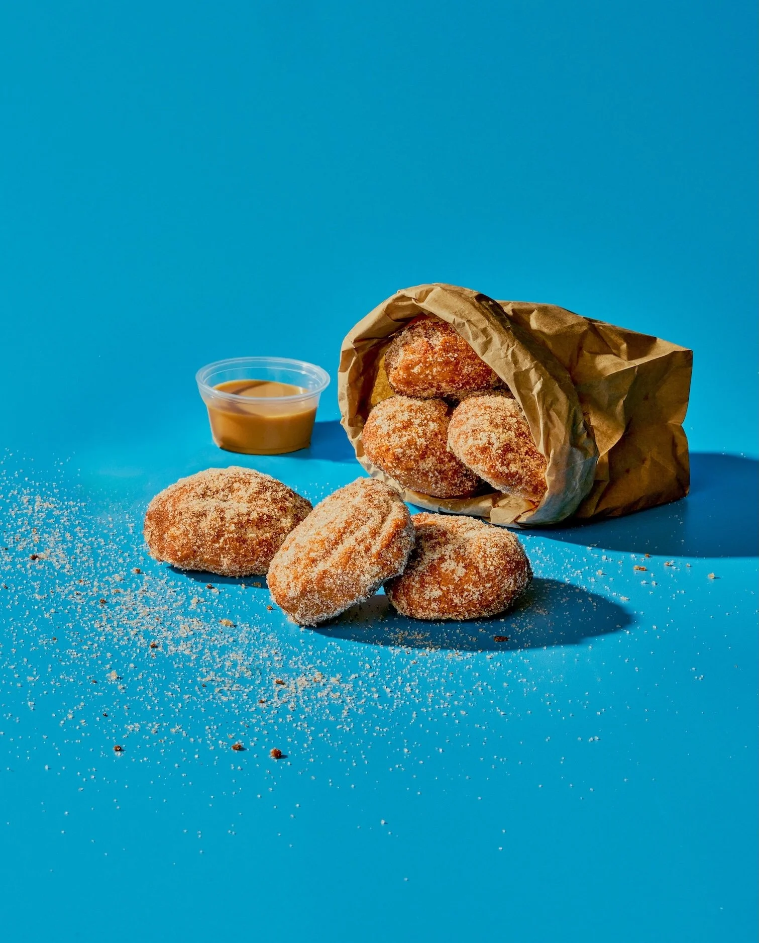Fried sugar-coated donuts spilling from a paper bag, with some on a blue surface, a small container of caramel sauce, and scattered sugar and chocolate sprinkles.
