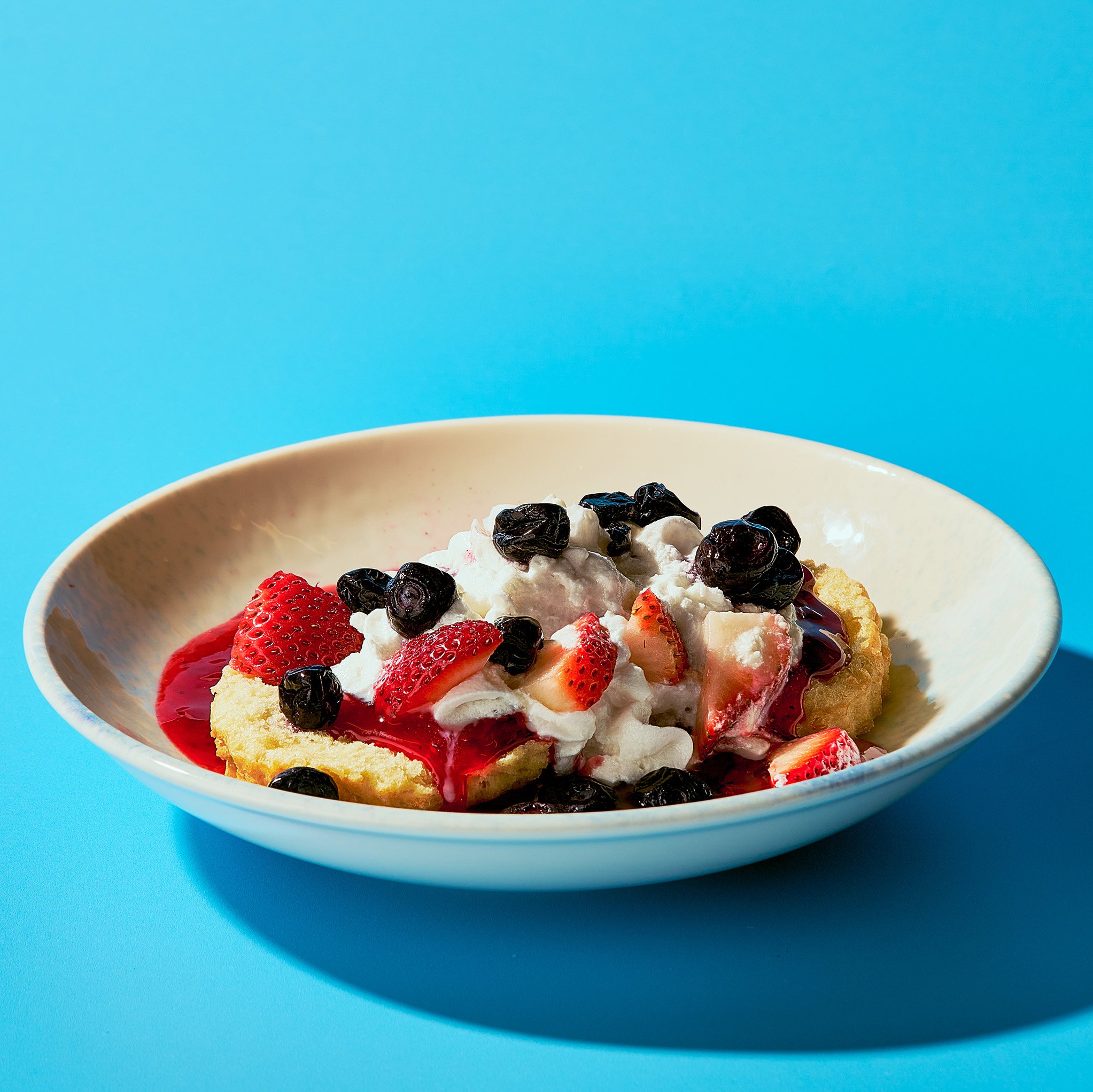 A bowl of dessert topped with strawberries, blueberries, whipped cream, and berry sauce on a blue background.