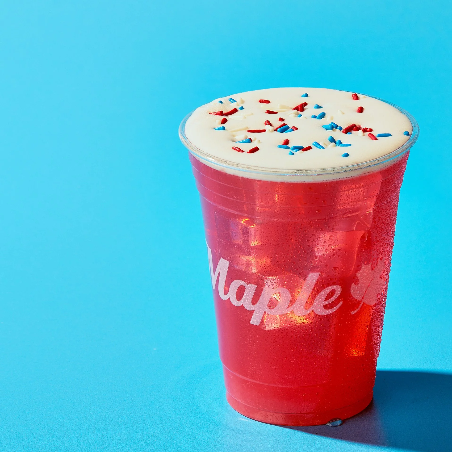 A red plastic cup filled with a creamy white beverage topped with red, white, and blue sprinkles, against a light blue background.