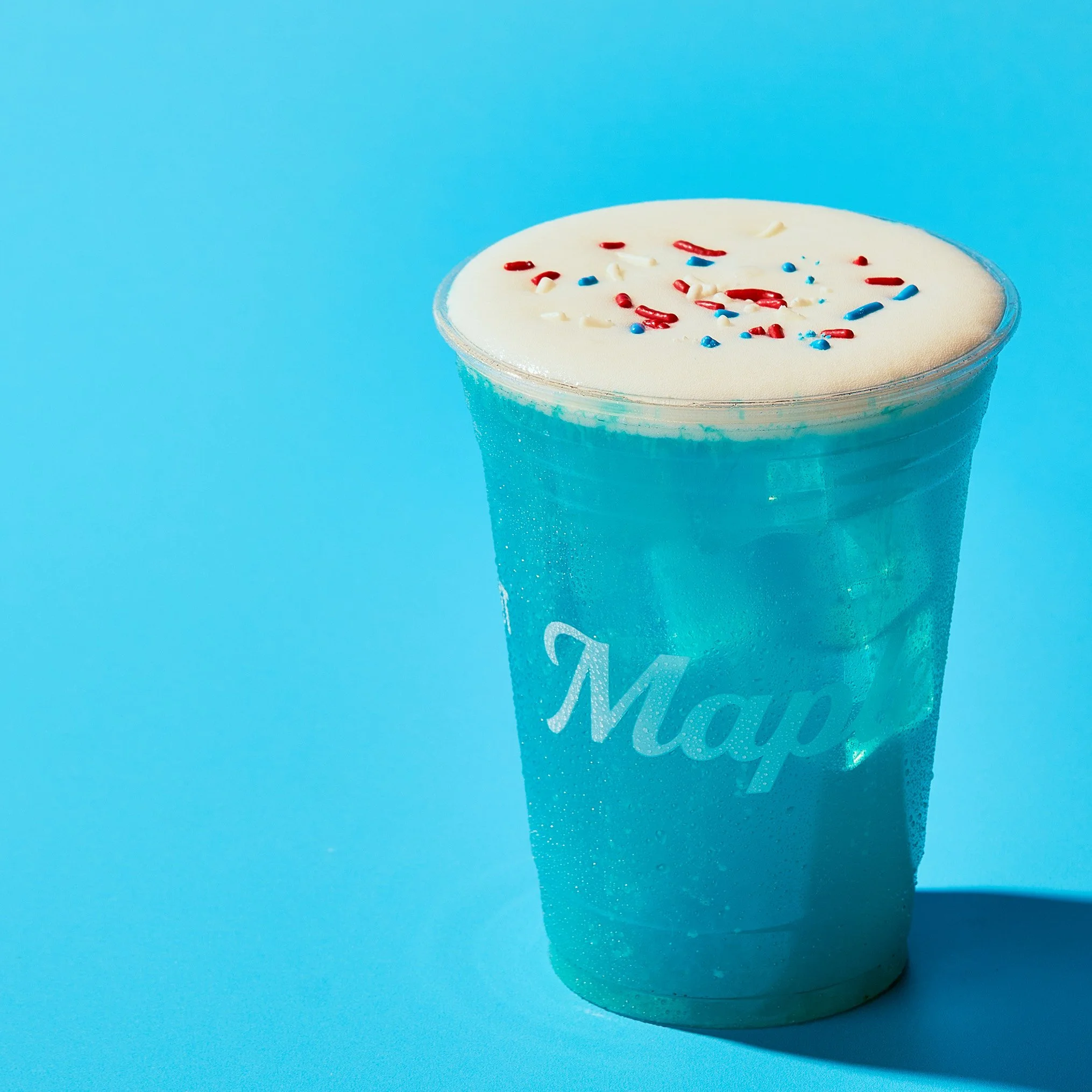 A cup of a creamy frozen dessert with red, white, and blue sprinkles on top against a bright blue background.
