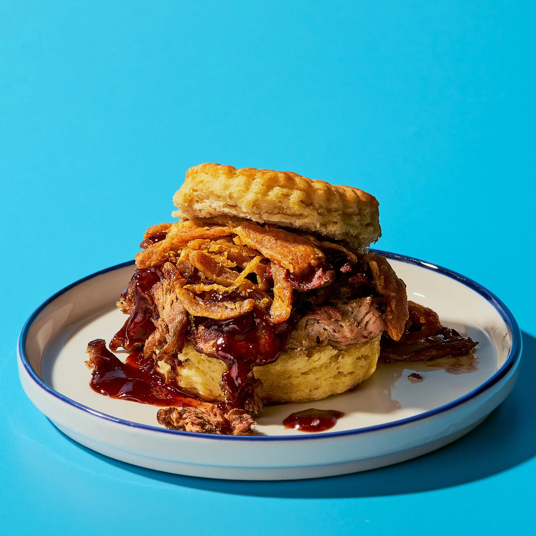 A plate of dessert with a bottom crust, topped with pulled pork covered in a dark barbecue sauce, a layer of crispy fried onions, and a piece of biscuit on top, set against a bright blue background.