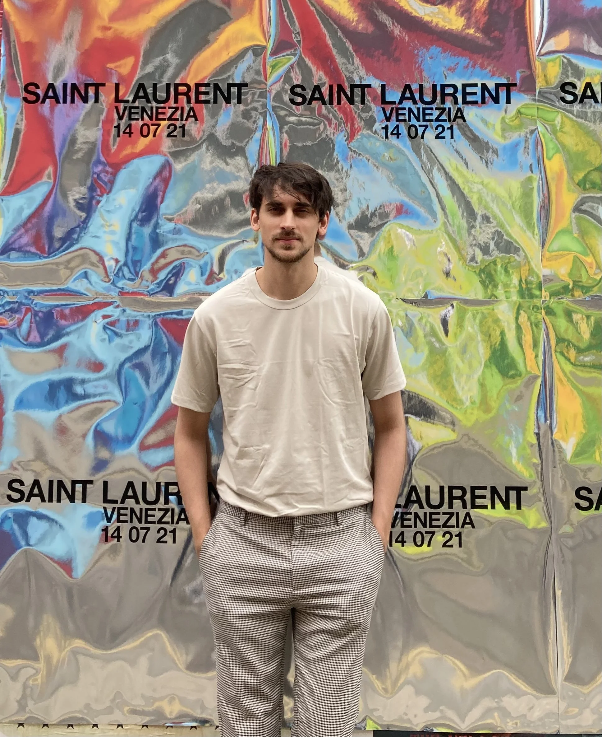 A young man standing in front of a colorful backdrop with the words 'Saint Laurent Venezia' and the date '14 07 21' printed on it. The backdrop has a metallic, abstract, multicolored design.