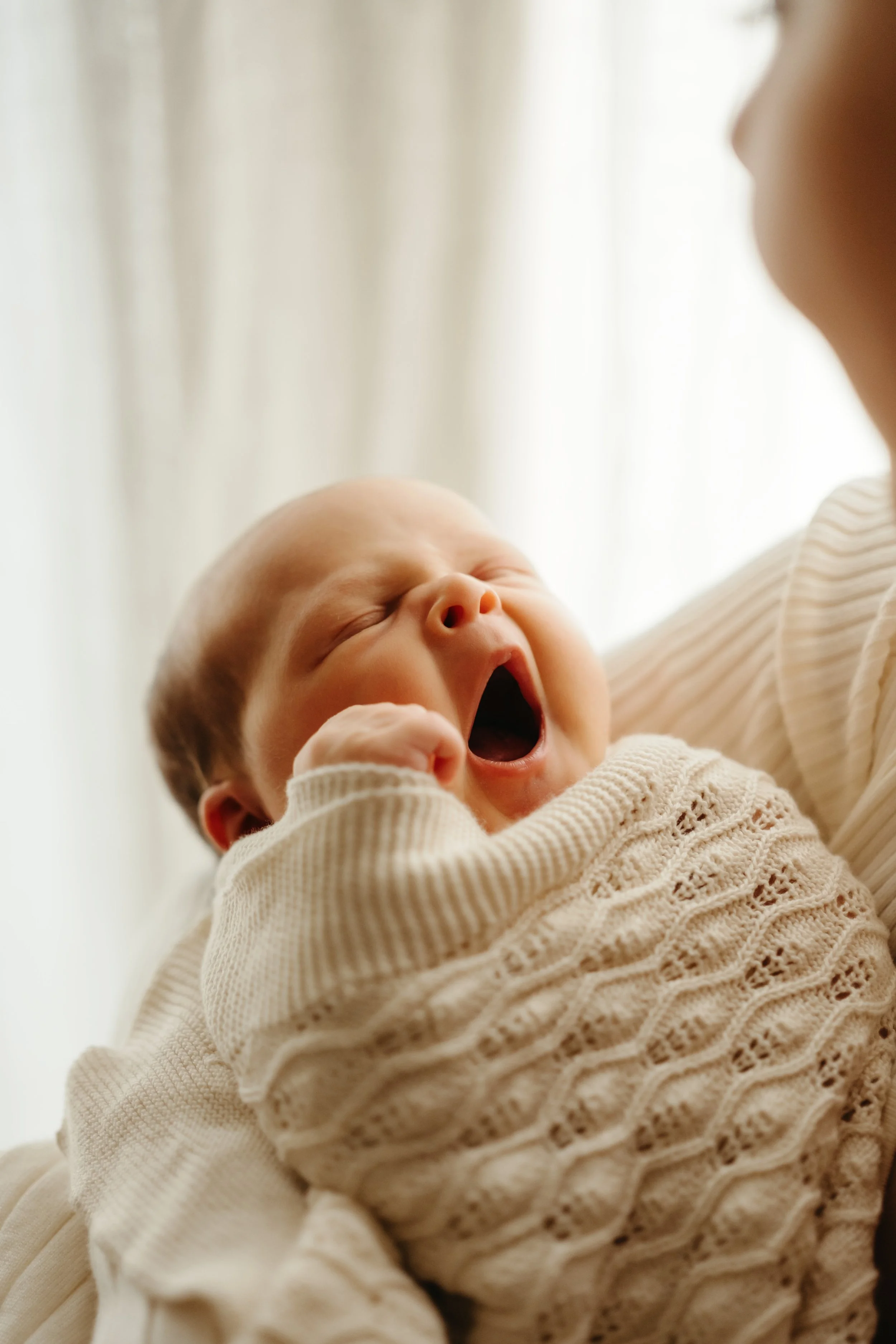 En baby som holder et fang, og yter en stor gjesp, med hvitt klær, mot et lyst bakteppe.