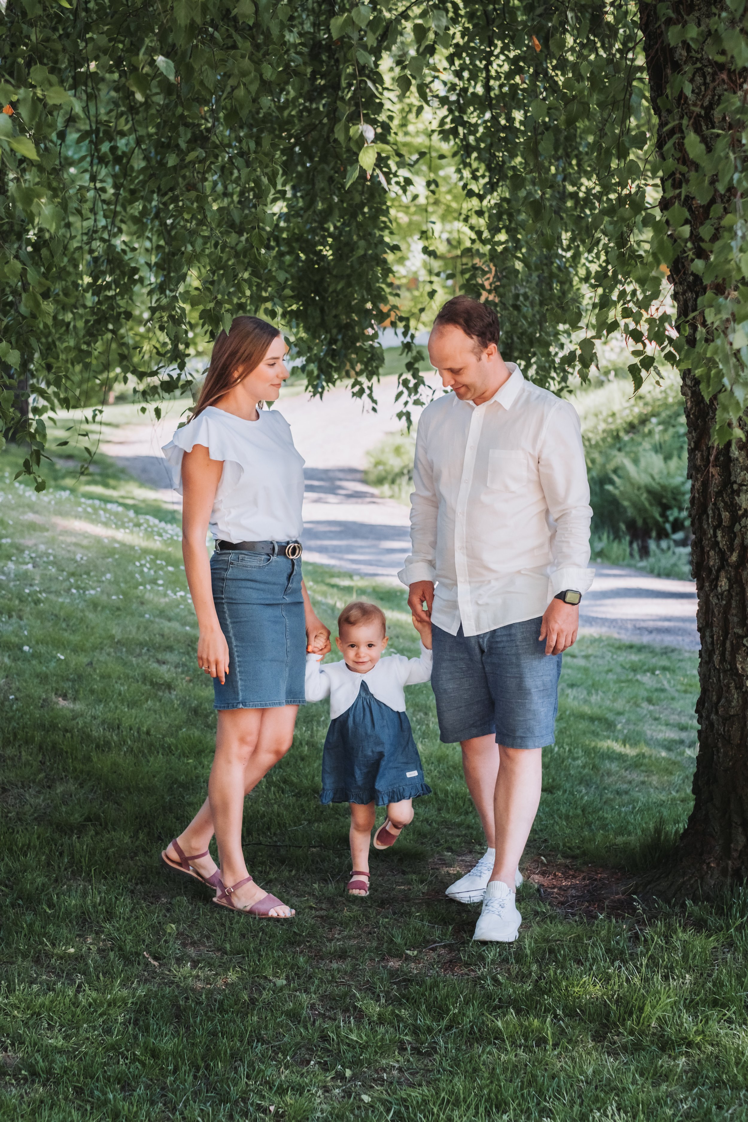 Family walking outdoors under a tree, holding hands with a young child. Parents are wearing casual summer outfits.