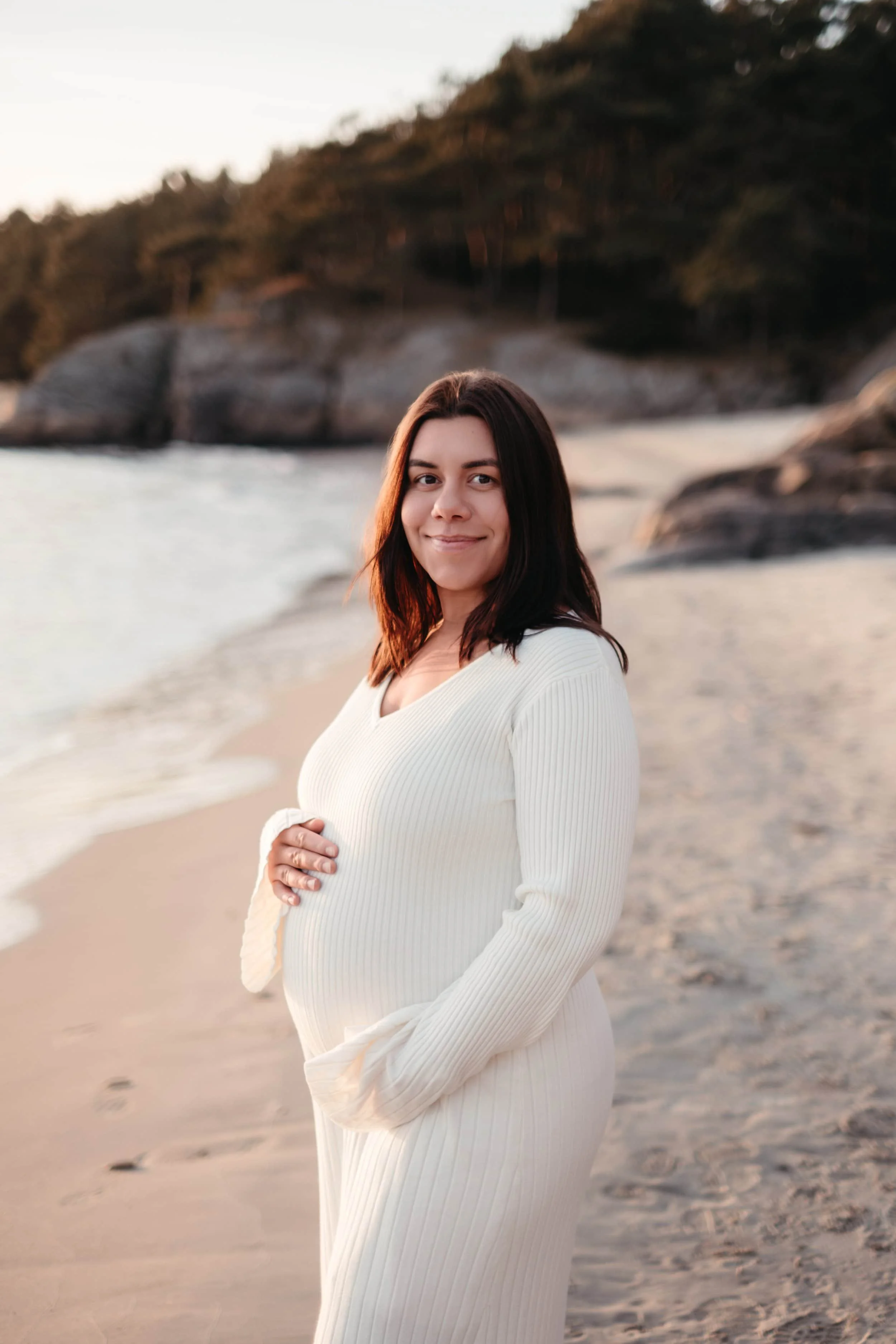 En gravid kvinne som står på en strand, smilende, med sjø og steiner i bakgrunnen under solnedgang.