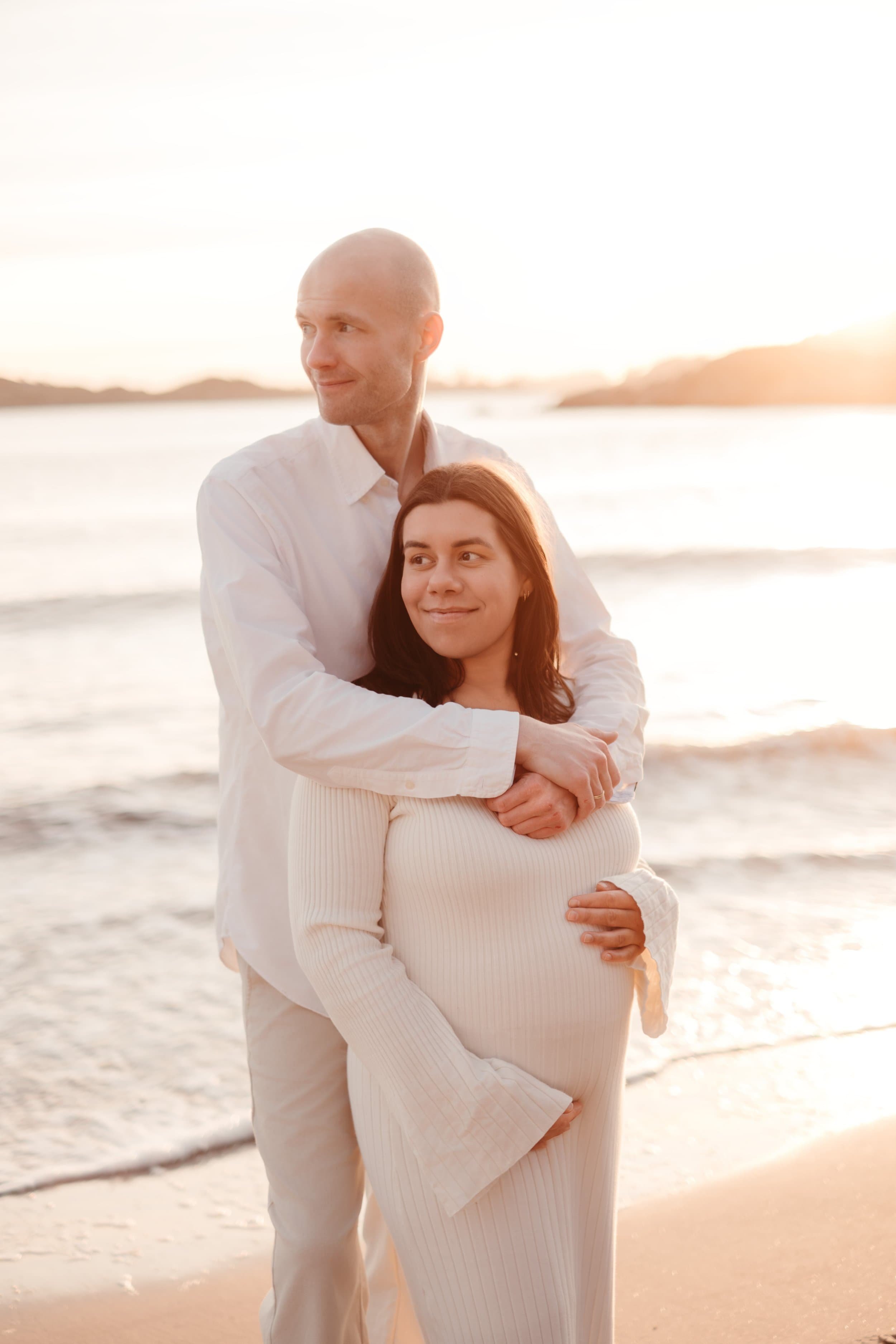 Et lykkelig par står nær sjøen ved solnedgang, med mannen som klemmer kvinnen rundt midjen, begge smiler.