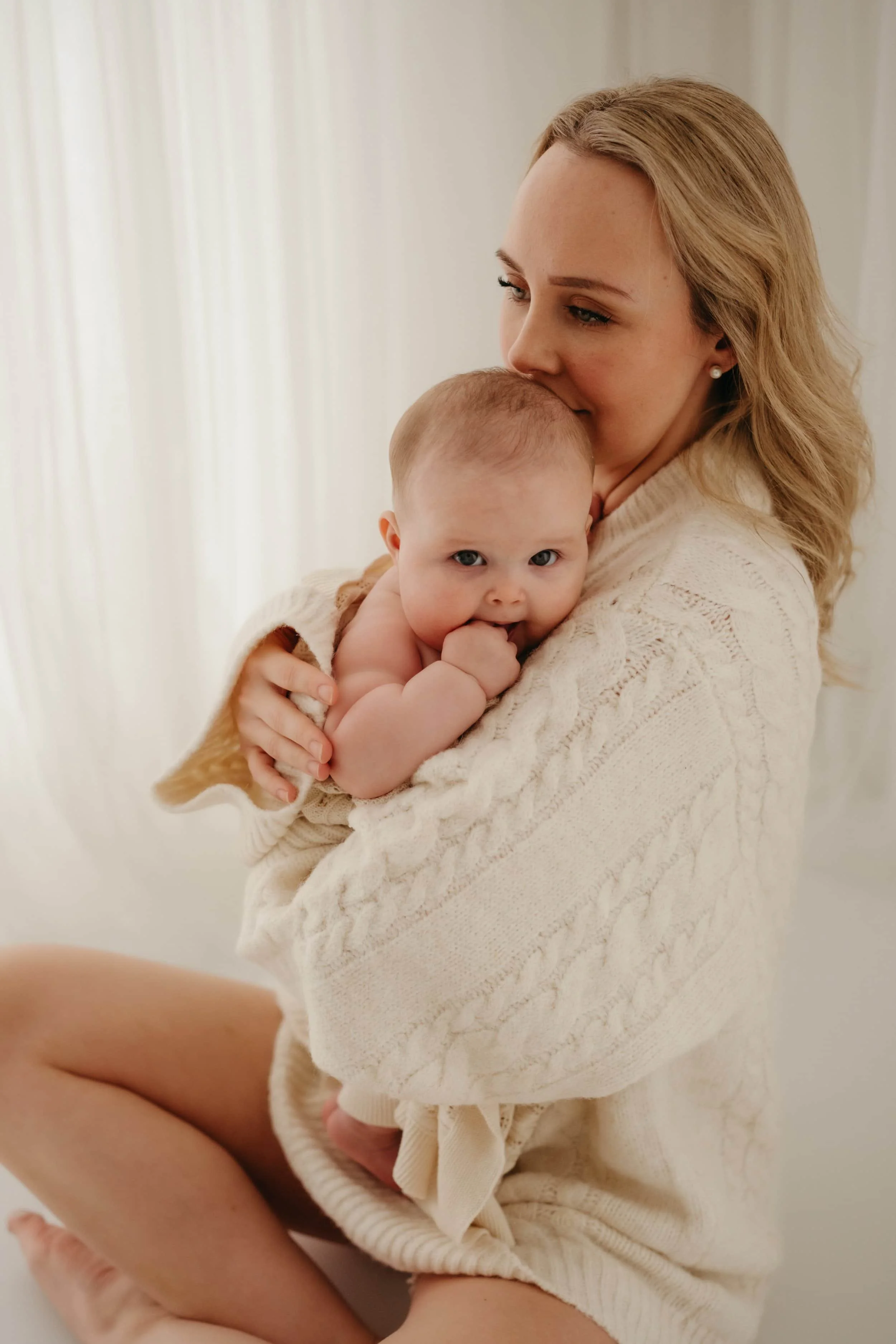 En kvinne holder en baby inntil brystet, begge har lyse hår og kvinnen smiler mykt.