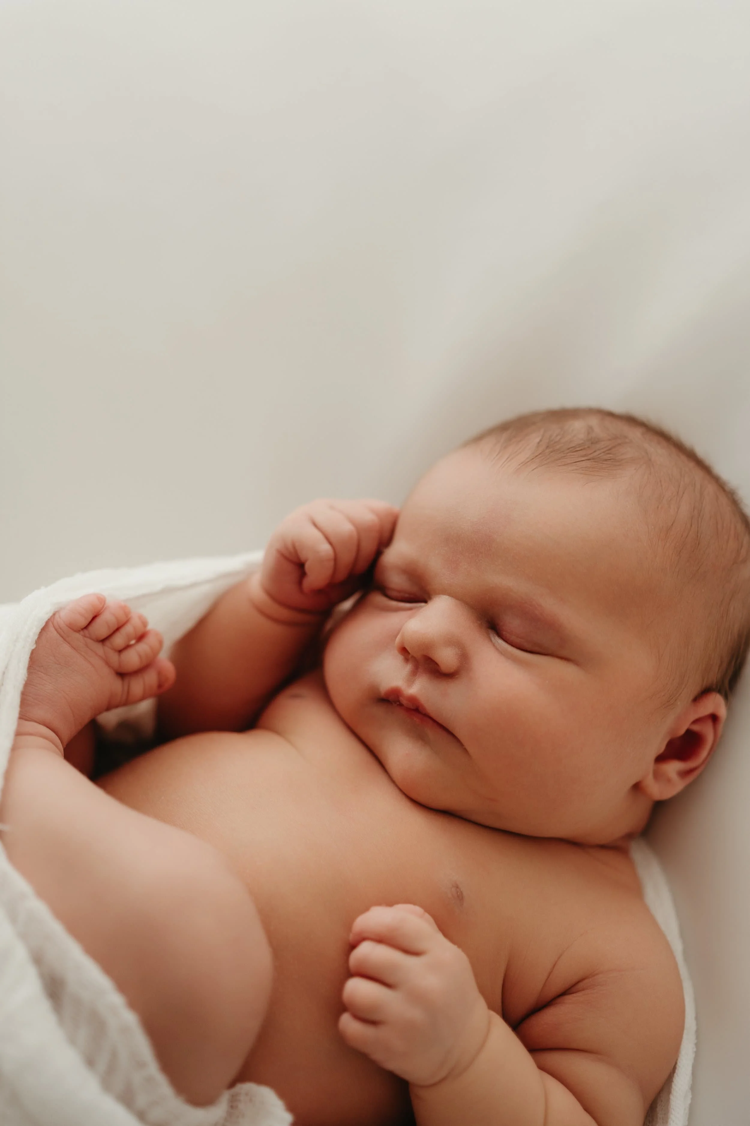 Nærbilde av en sovende baby med lukkede øyne og liten hånd ved ansiktet, delvis dekket av et hvitt teppe.