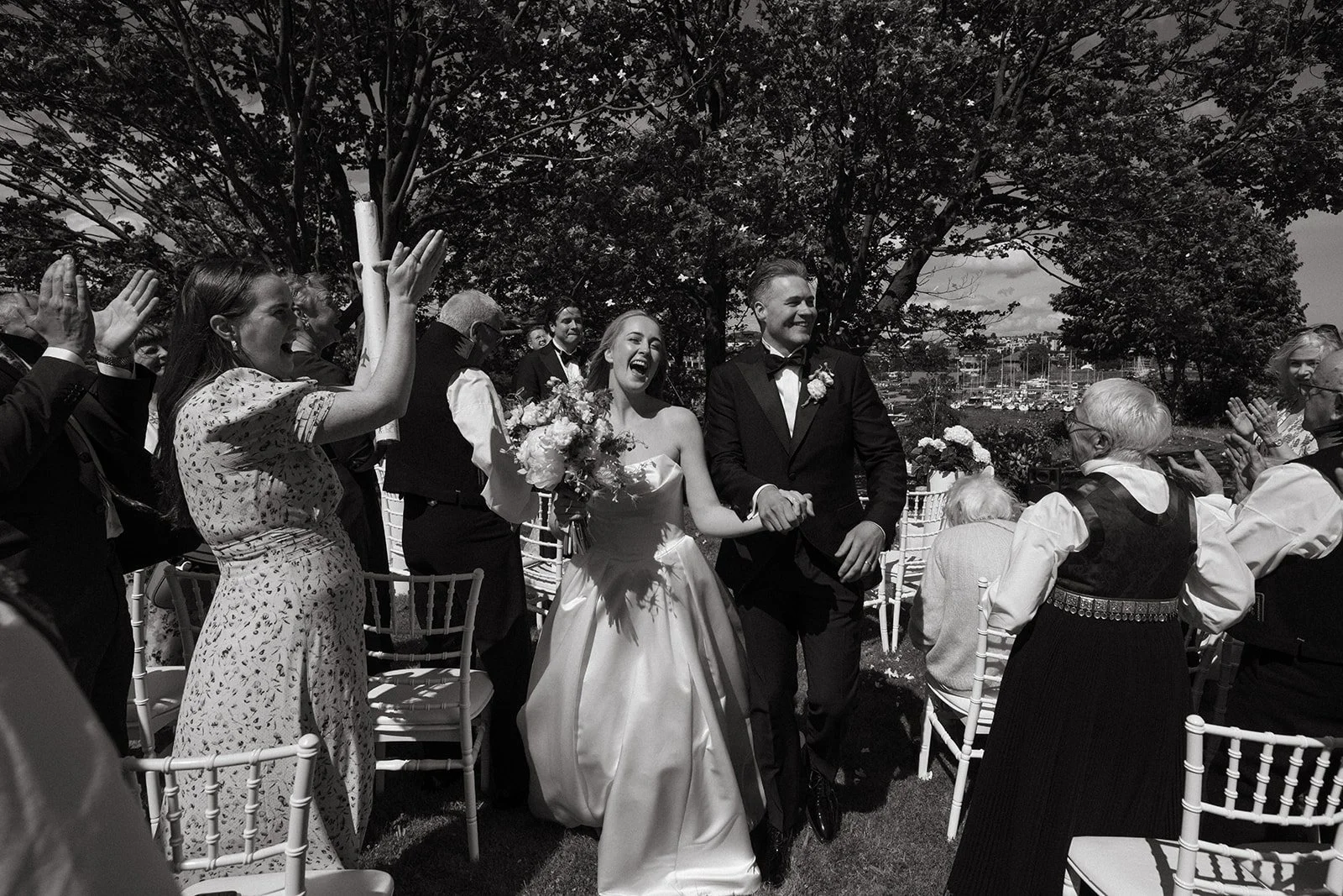 En brudepar går ned Midtgangen i et utendørs bryllup, omgitt av gjester som applauderer, med en marina med båter i bakgrunnen.
