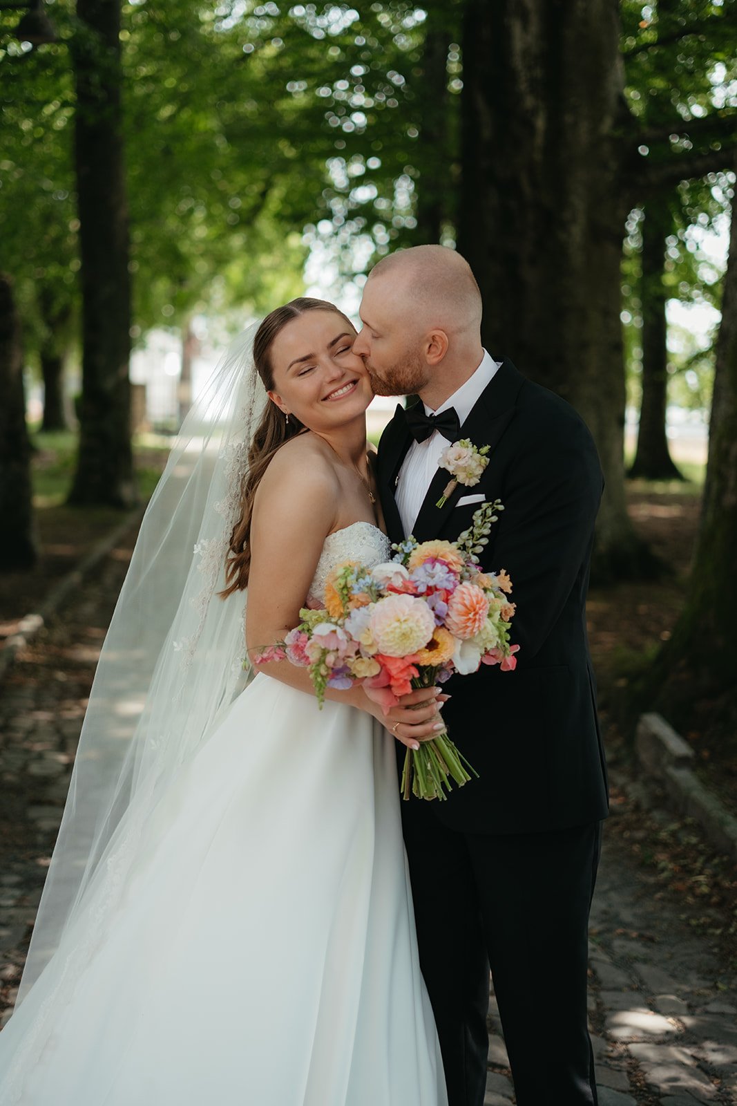Et brudepar i et skogsområde, hvor bruden holder en blomsterbukett og mannen kiler henne på kinnet, begge smilende.