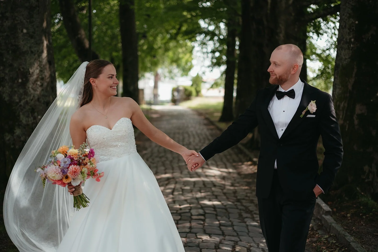 Par i bryllupsantrekk holder hender og smiler til hverandre på en stiene i en park med store trær.