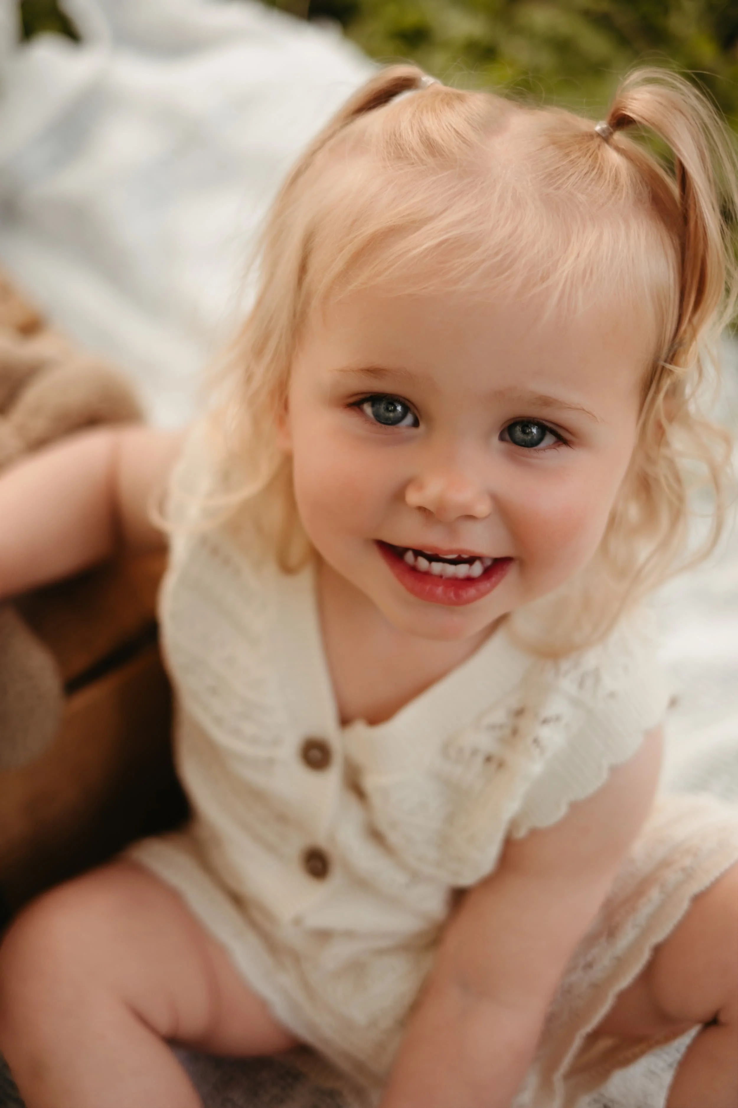 Liten jente med blond, krøllet hår og blå øyne smiler, iført hvit genser, utendørs.