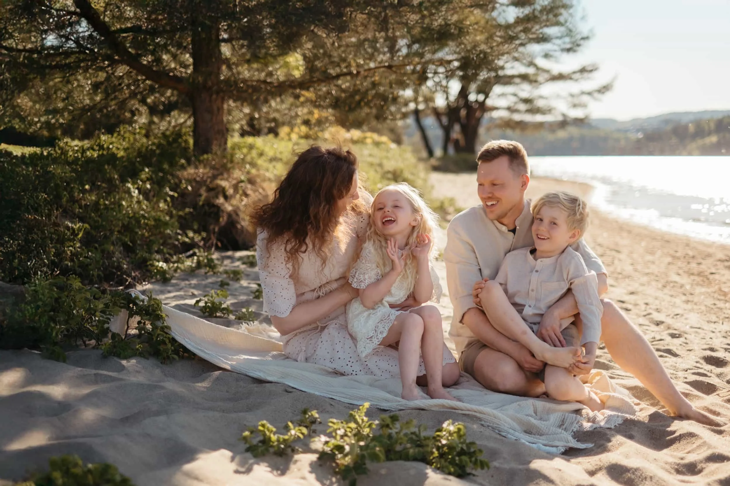 En familie som har det gøy sammen på stranden, ler og smiler mens de sitter på sanden under trær med utsikt over vannet.
