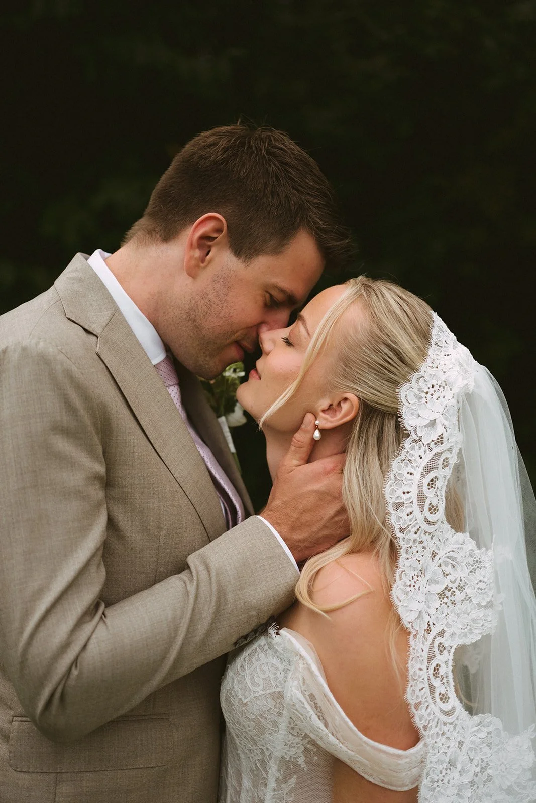 Et bryllupsbilde av et par som kysser med pannen mot pannen, kledd i bryllupsantrekk. Mannen har på seg en beige dress, og kvinnen har på seg en hvit brudekjole med blondedetaljer og skrått sidepannel. De står utendørs med mørk, grønn bakgrunn.