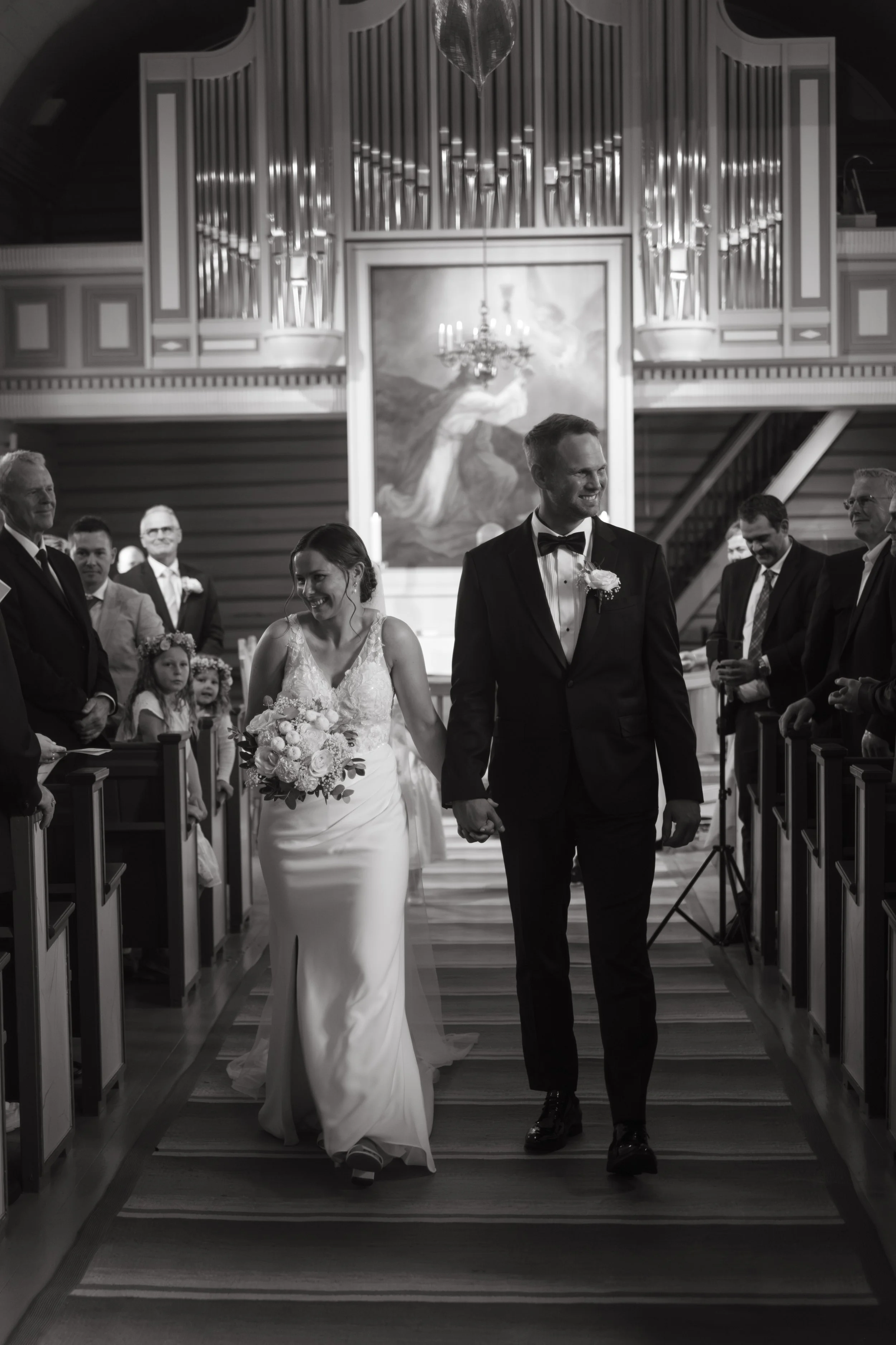 Et brudepar går ned midtgangen i en kirke, omgitt av familie og venner, under et bryllup, i svart-hvitt, med folk som smiler.